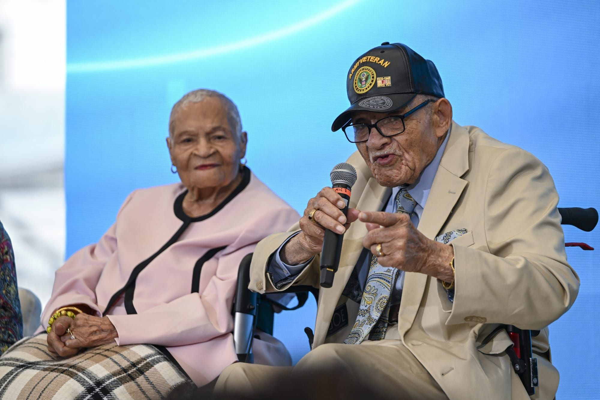 color photograph of two elderly Black people sitting on stage. the man on the right holds a microphone as he speaks