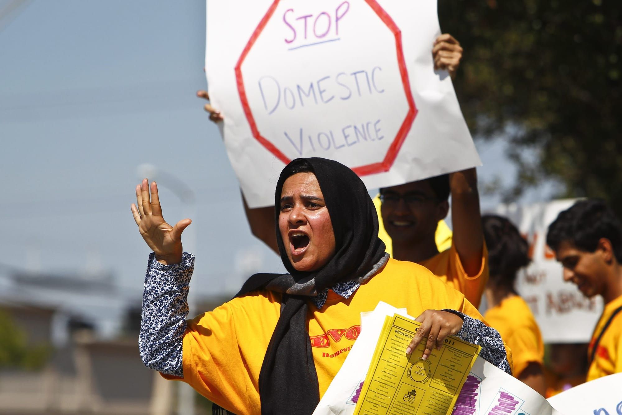 color photograph of an outdoor protest. a woman in a black head scarf wearing a yellow shirt stands in the foreground shoutin