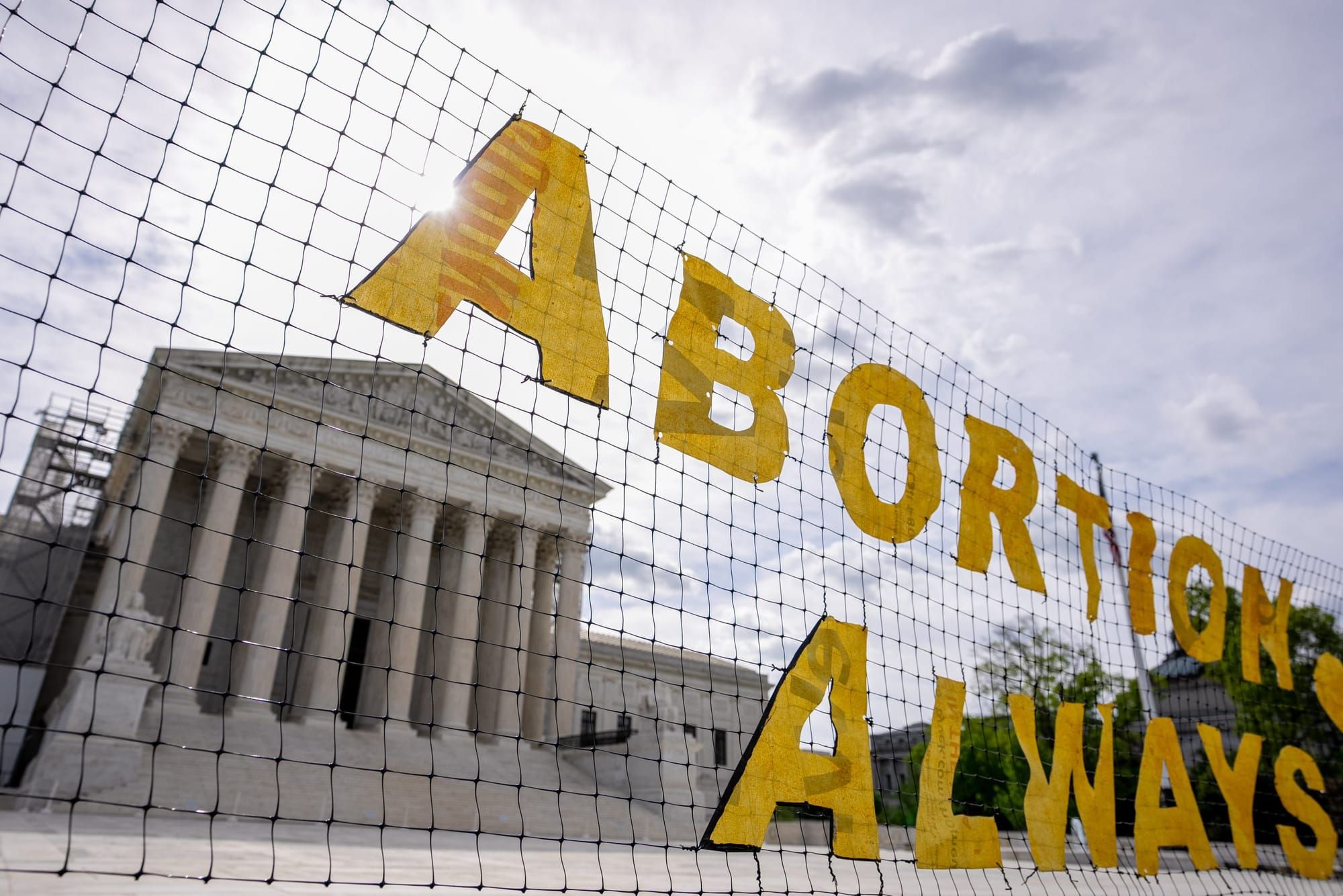 color photograph of a net fence in front of the Supreme Court building. Yellow block letters reading Abortion Always are atta