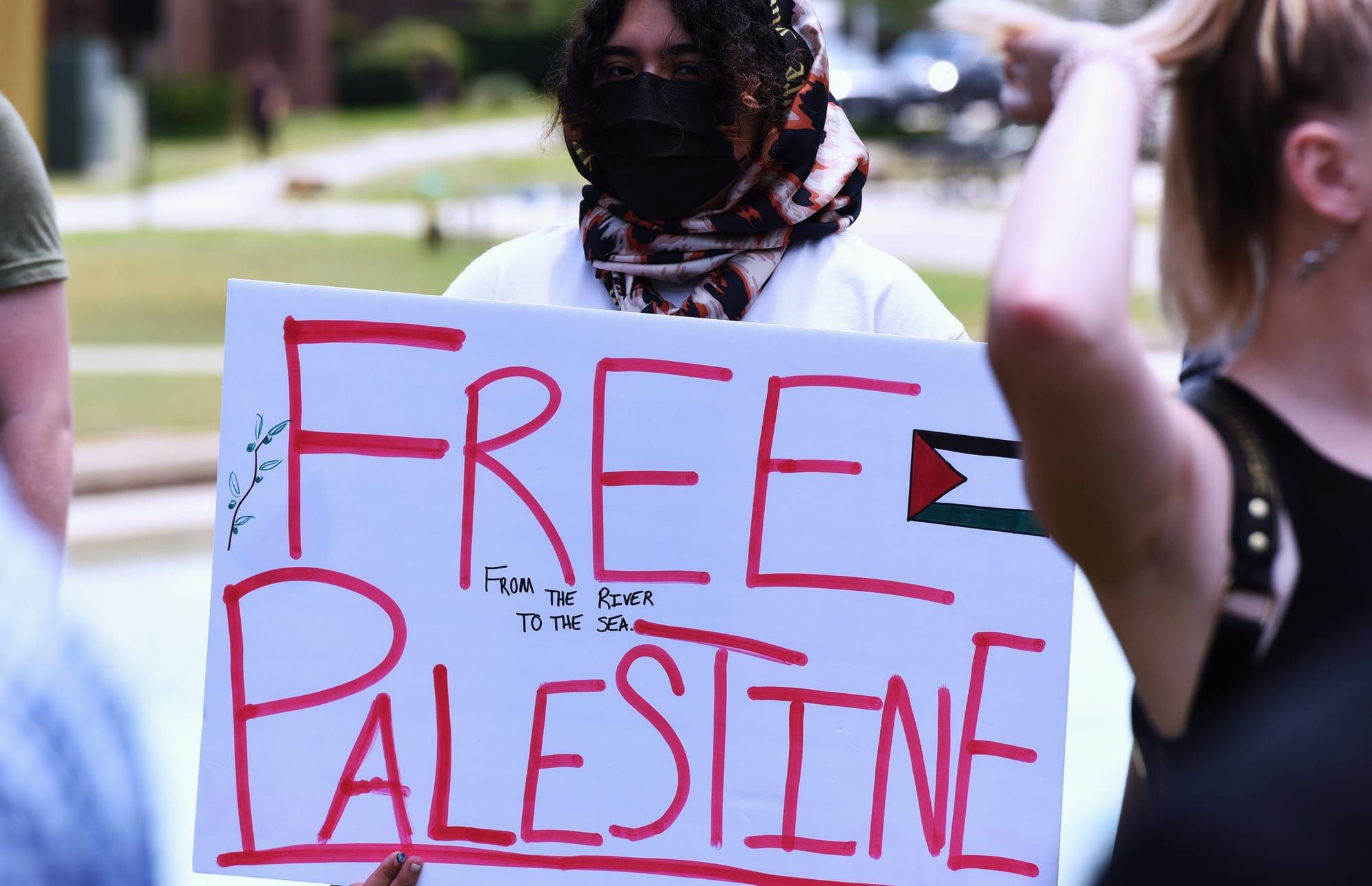 color photograph of a protester holding up a white sign with red handwritten text that reads "free palestine from the river t