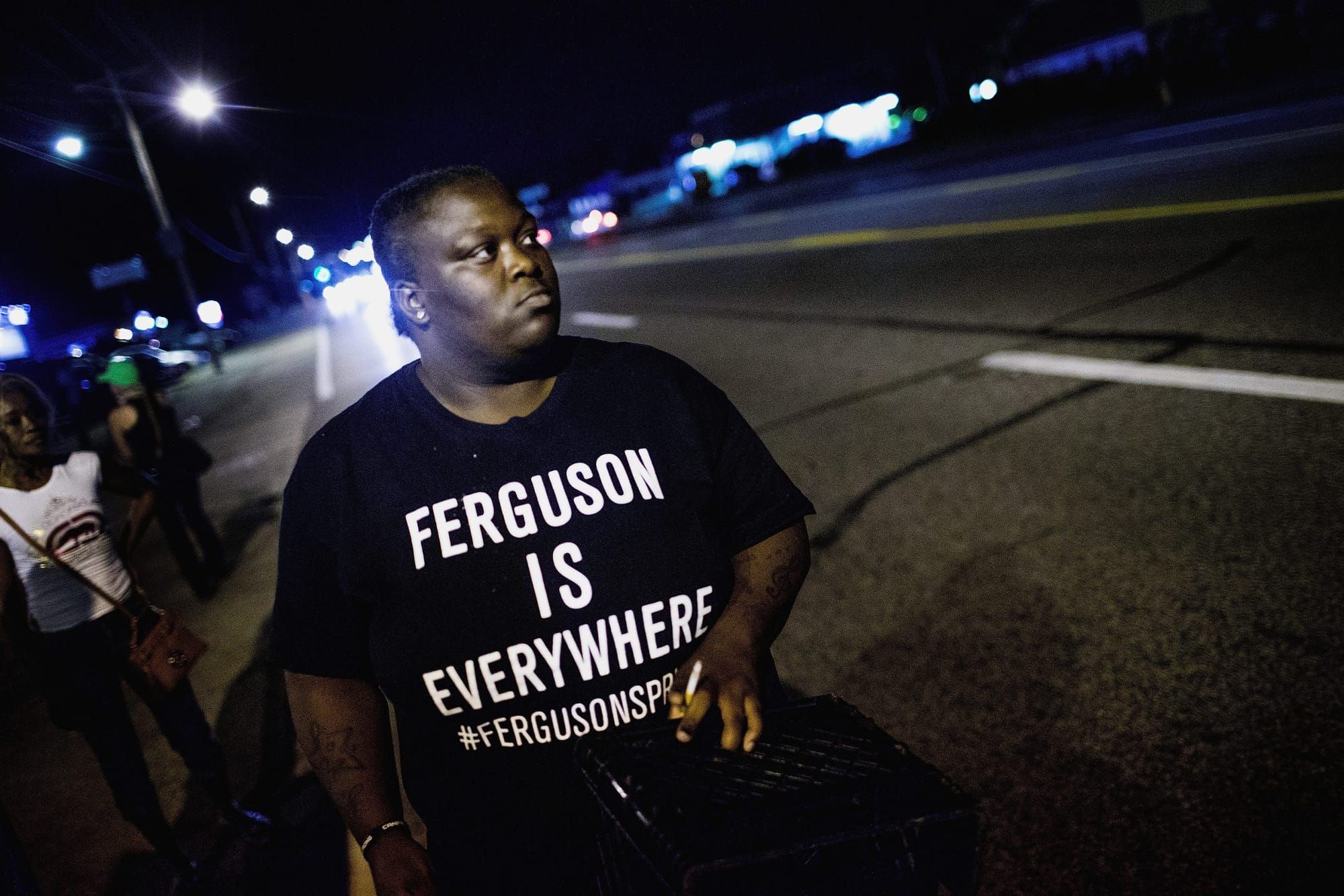 color photograph at night of a Black demonstrator wearing a "Ferguson is everywhere" shirt walking in the middle of a multi-l