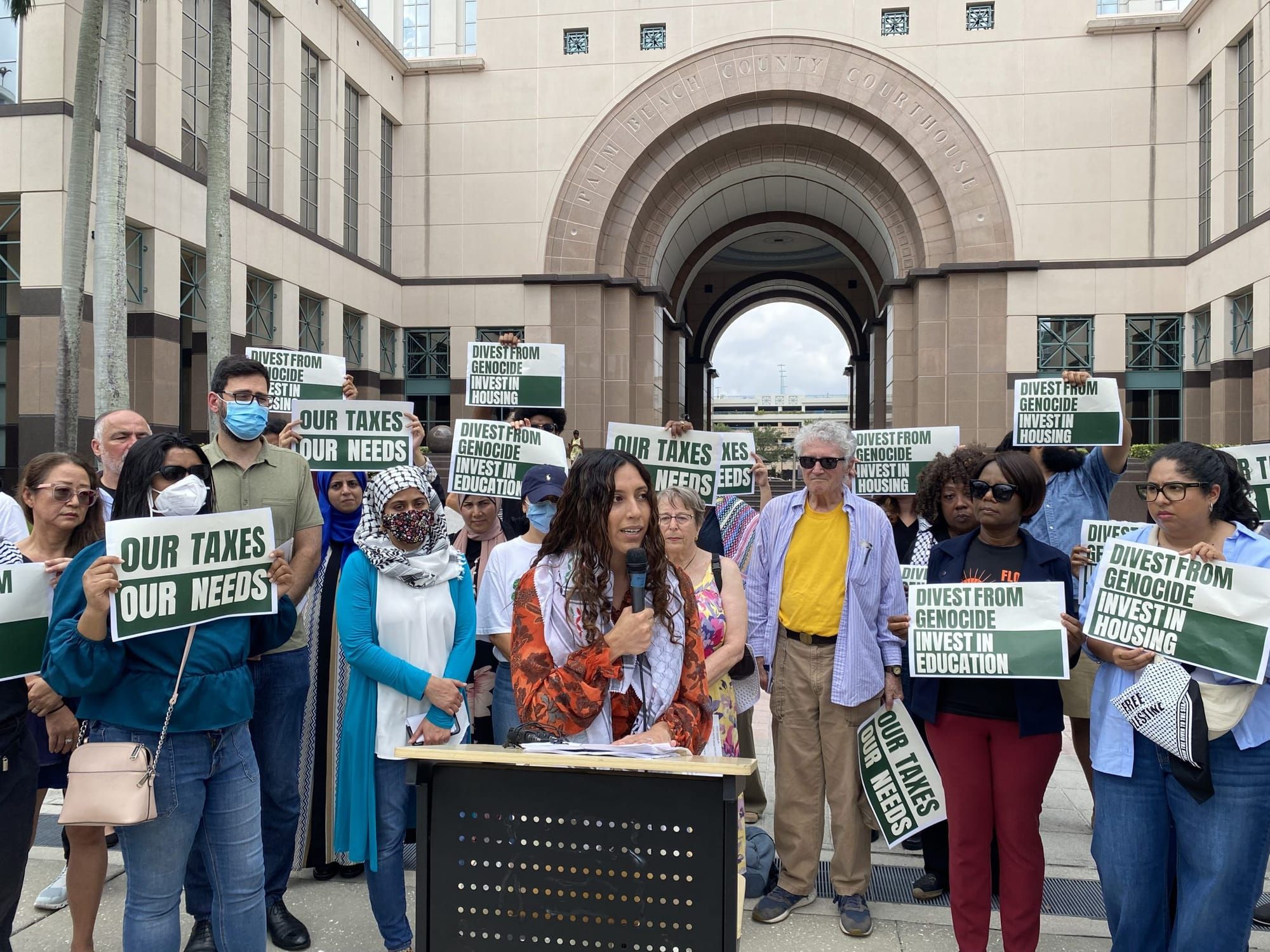 color photograph of an outdoor press conference. people hold signs reading "divest from genocide invest in housing" and "our