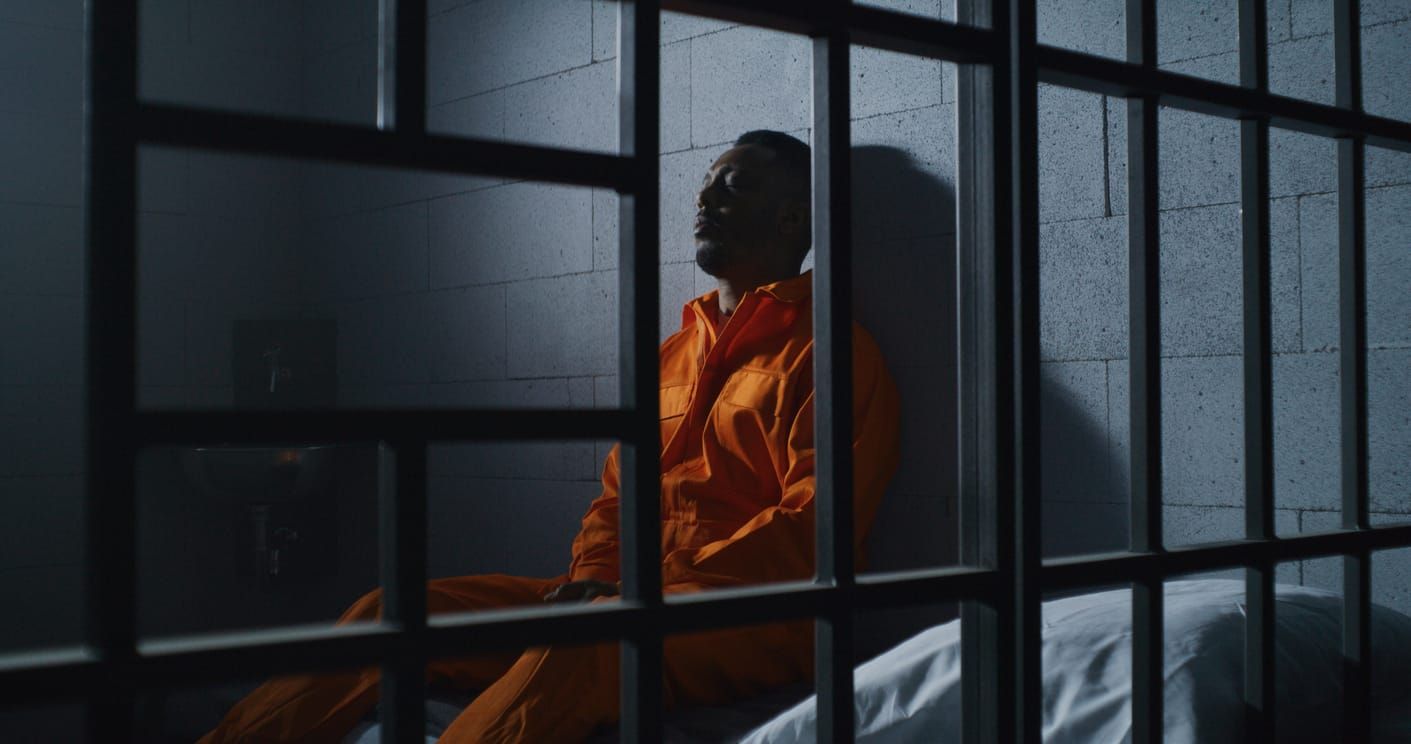 color photograph of a Black man in an orange jumpsuit sitting behind metal bars
