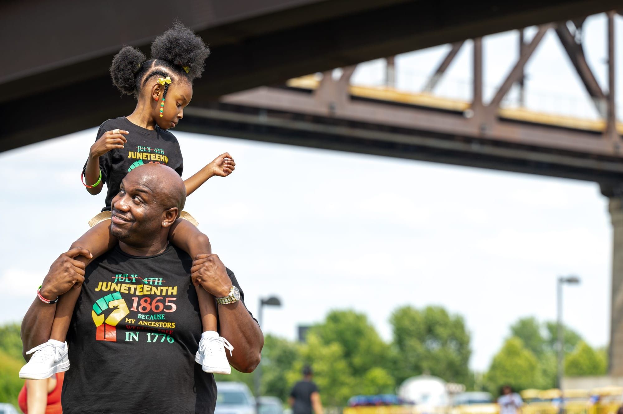 color photograph of a young Black girl sitting on a man's shoulders at a Juneteenth celebration
