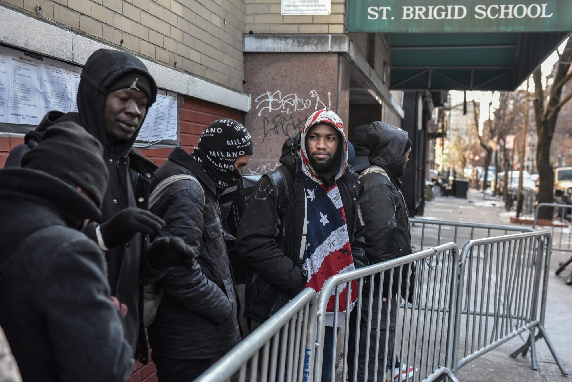 color photograph of Black men in winter coats lined up outside a stone building with a green awning that reads "St. Brigid Sc