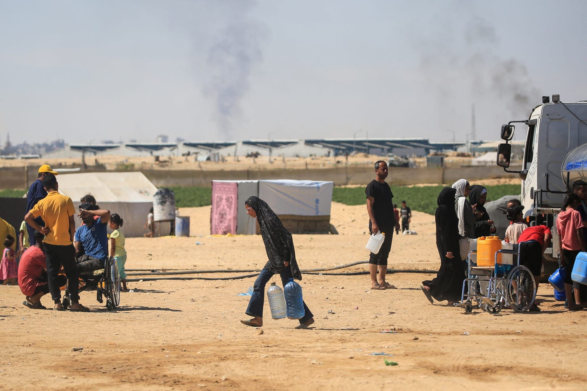 color photograph of a woman in a Palestinian camp carrying two large jugs of water in front of a skyline dotted with rising s