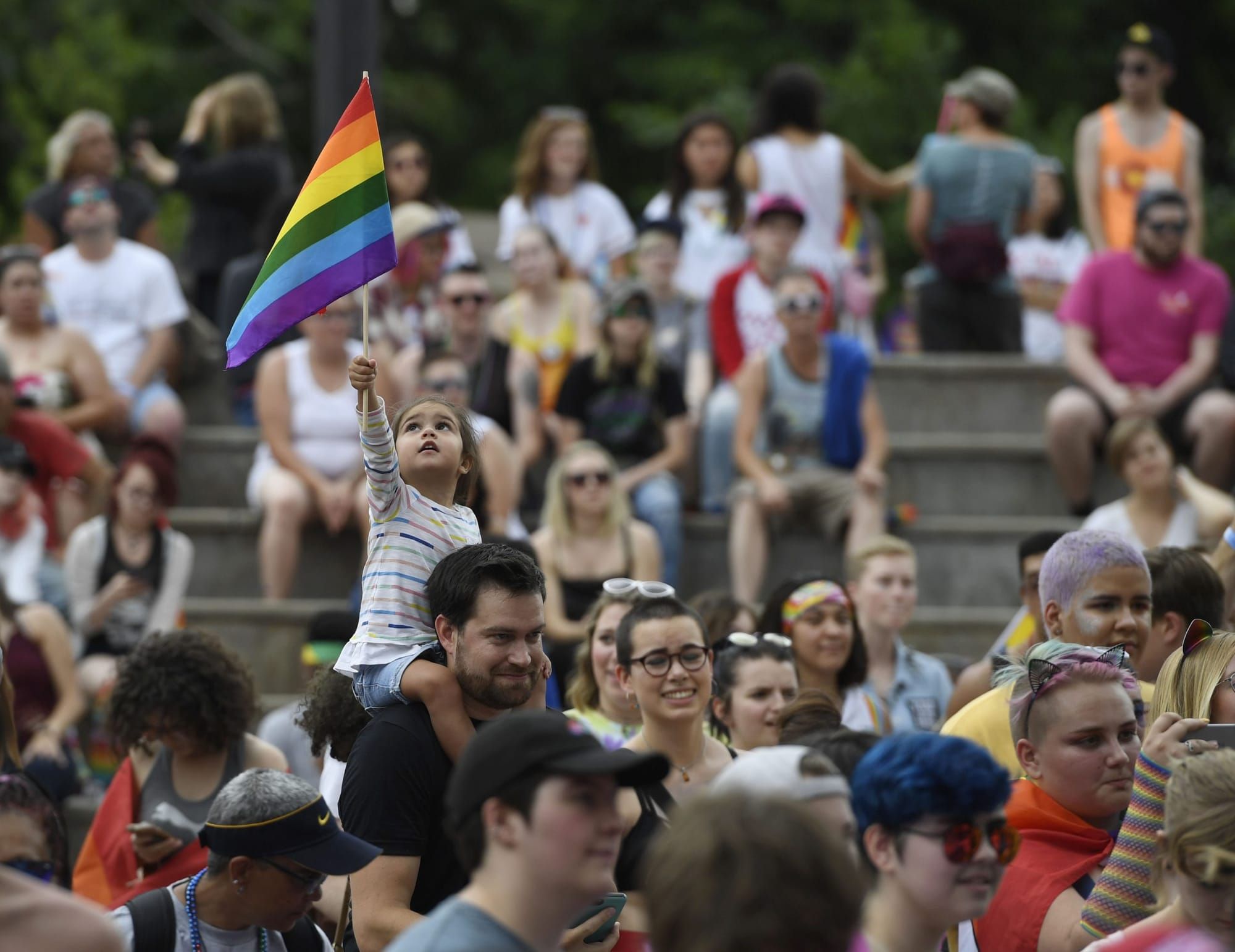 color photograph of an outdoor Pride celebration. A young child sits on their father's shoulders and waves a Pride flag