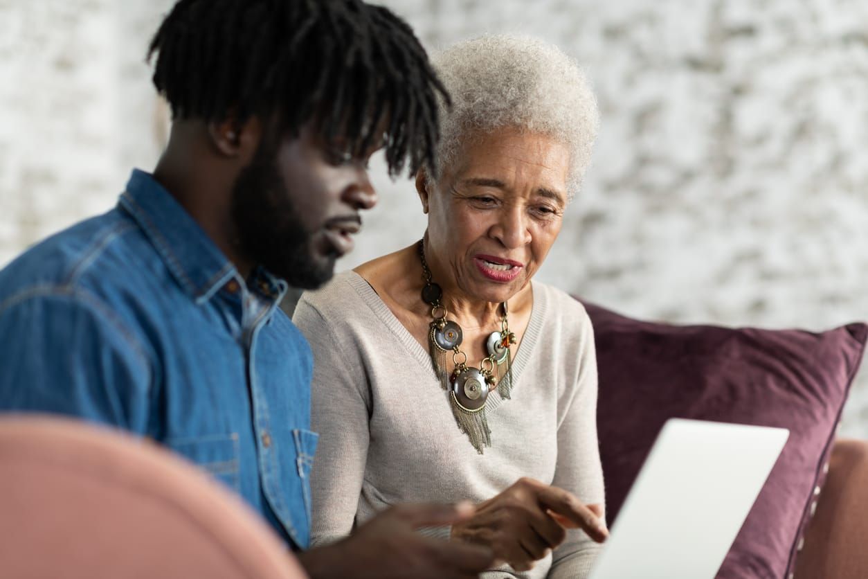 stock color photo of a young Black man sitting next to his grandmother teaching her how to use a tablet computer