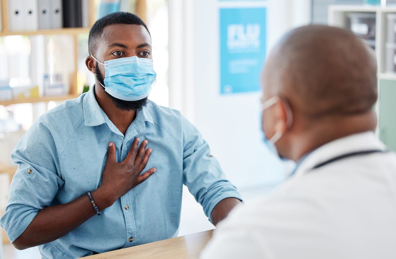 color stock photo of a Black man in a blue button-up shirt wearing a medical mask talking to a doctor whose back is to the ca