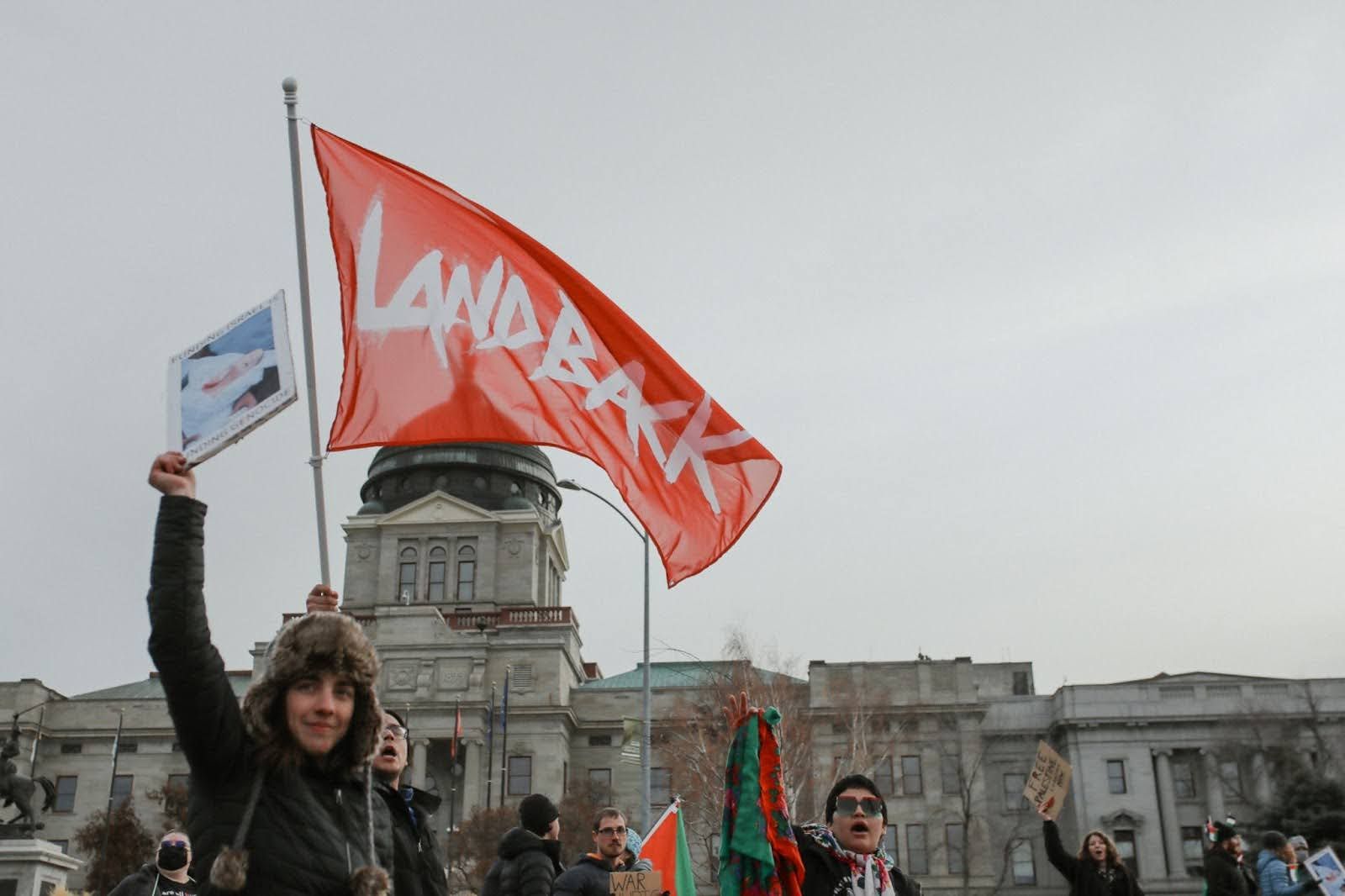 color photograph of an outdoor protest in front of a state capitol building. a person in the foreground waves a red "LANDBACK