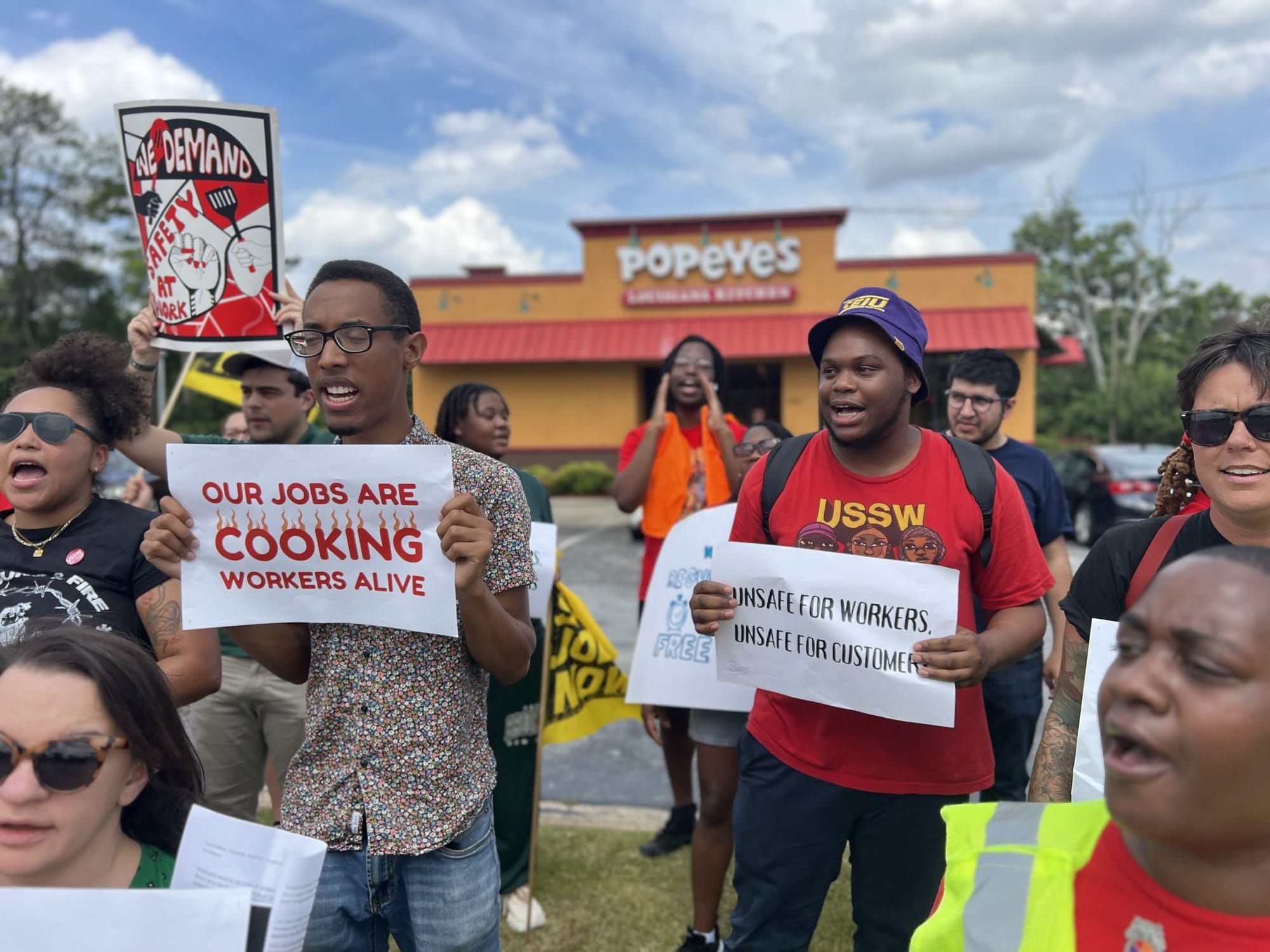 color photograph of an outdoor protest in front of a Popeyes. people hold signs reading "our jobs are cooking workers alive"