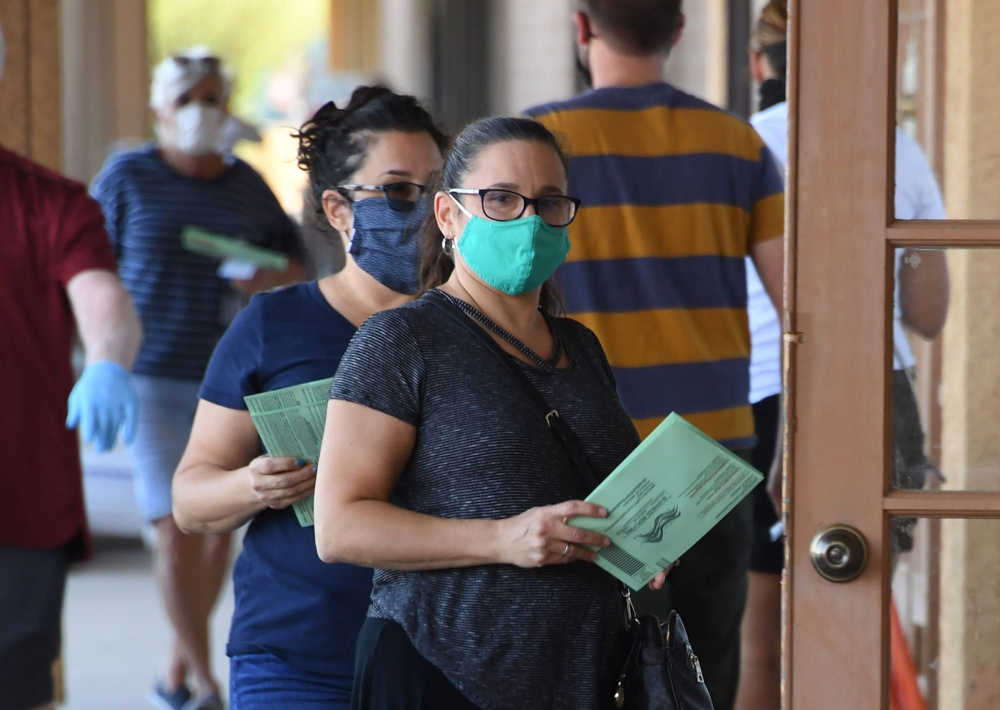 color outdoor photograph of masked Latinas standing in line and holding mail-in ballots