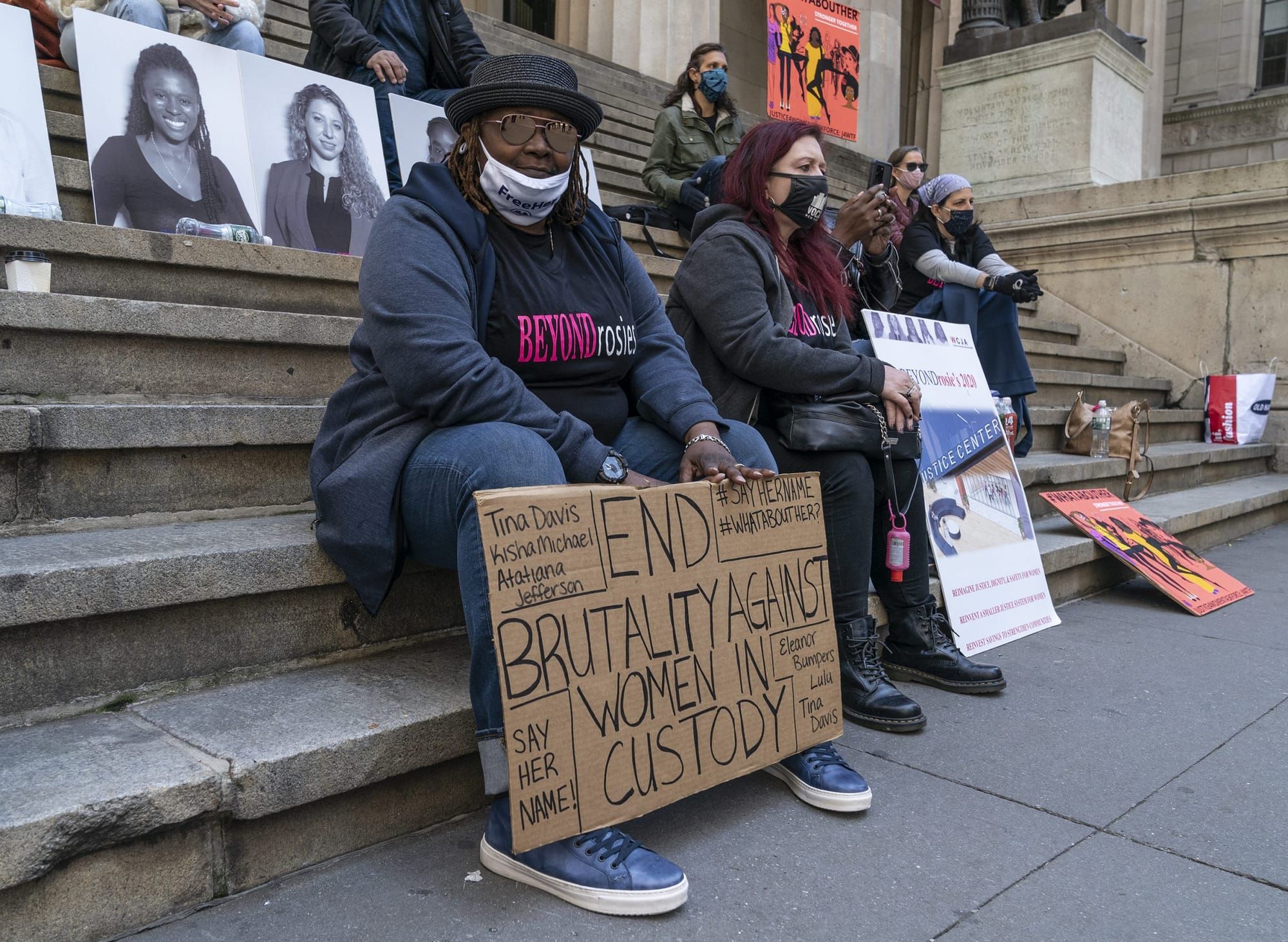 color photograph of Black women sitting on the steps of a government building. the woman in the center holds a cardboard sign