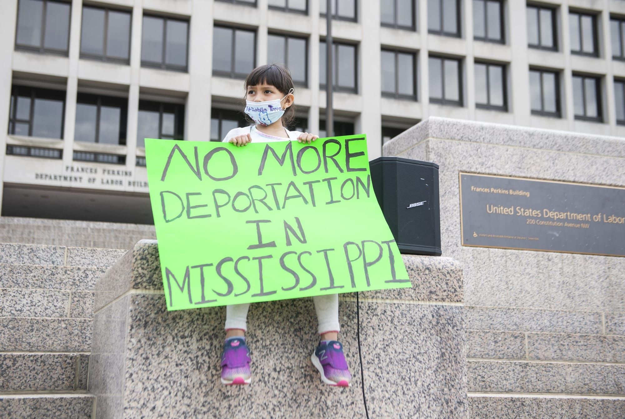 color photograph of a young girl wearing a mask sitting on a ledge holding a green poster that reads "no more deportations in
