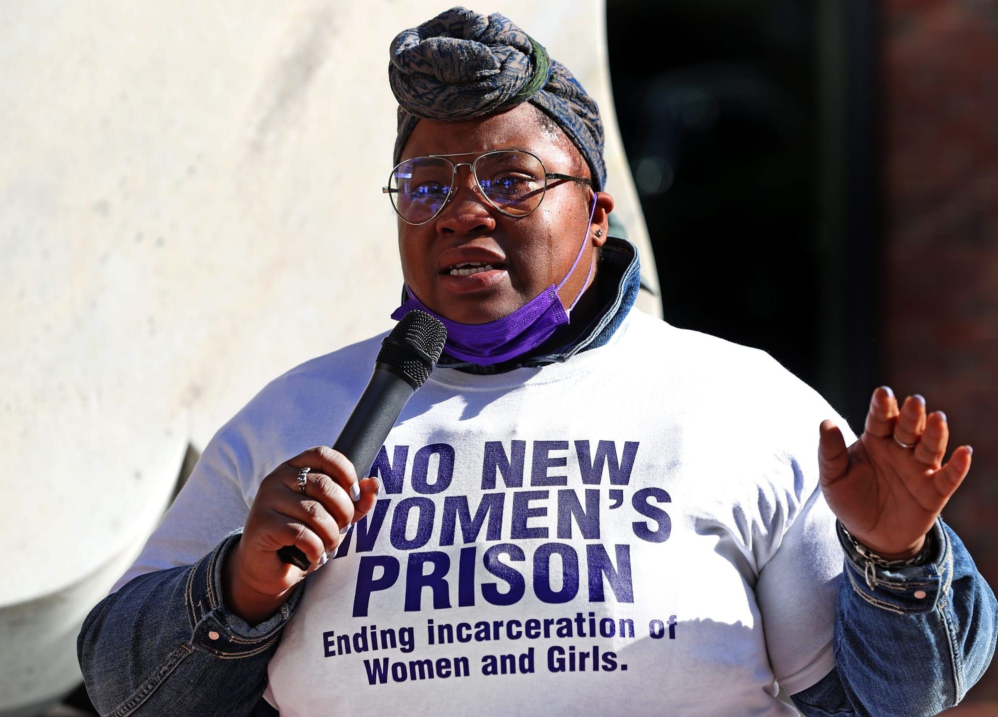 color photograph of a Black woman holding a microphone and wearing a white T-shirt that reads "no new women's prisons"