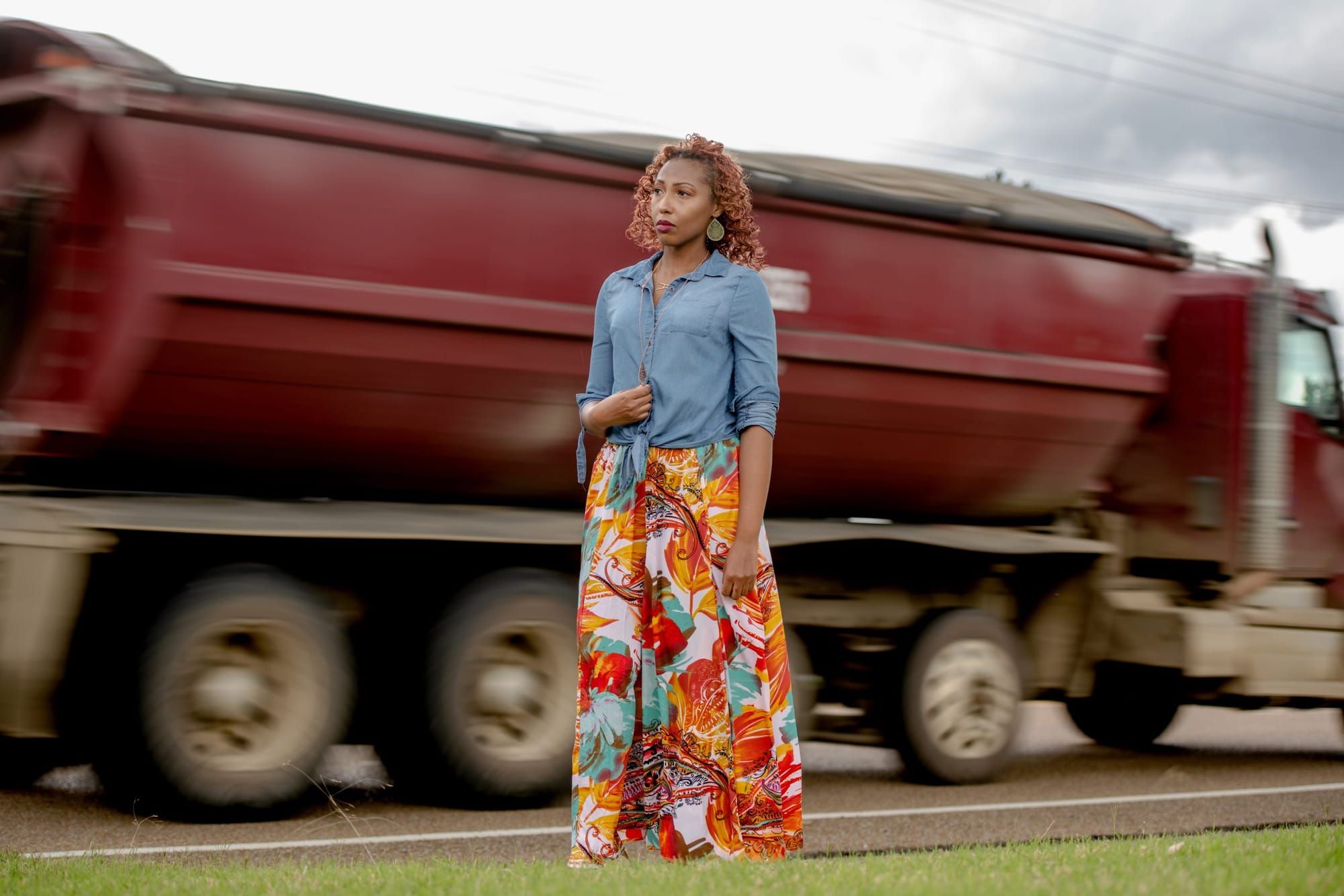 color photograph of a Black woman in a long bright skirt standing by the side of a road as a coal truck speeds past