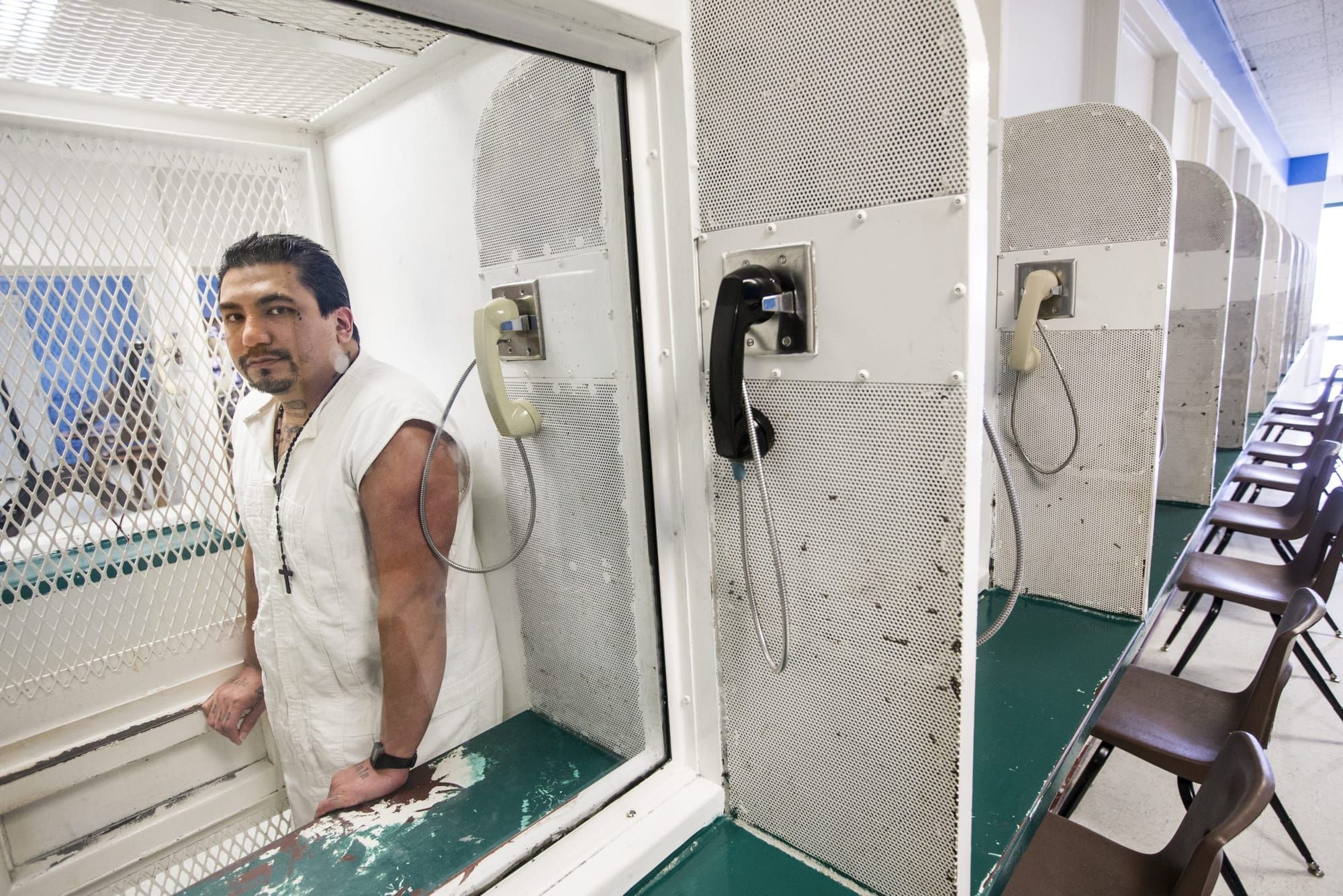 color photograph of a man with tattoos on his arm wearing a white prison jumpsuit. he stands behind a glass barrier with a ph