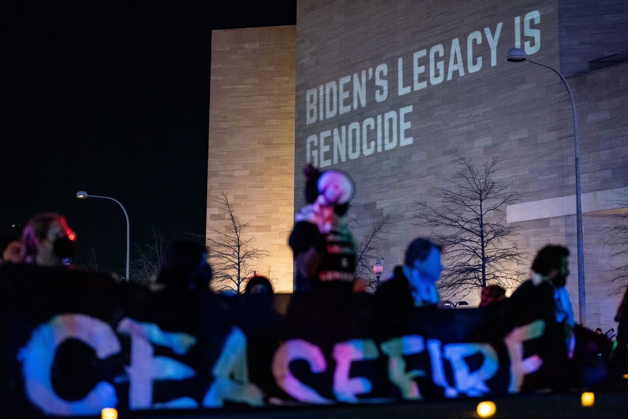 color photograph of an outdoor nighttime protest. people hold a banner reading "ceasefire" in the foreground, and the words "
