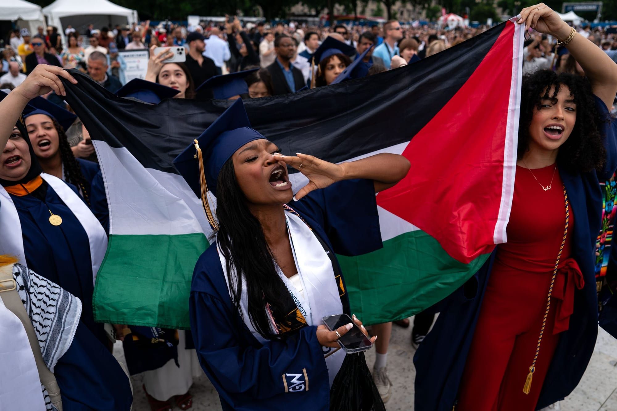 color photograph of a Black woman in a graduation cap and gown calling out to others as she stands in front of a Palestinian