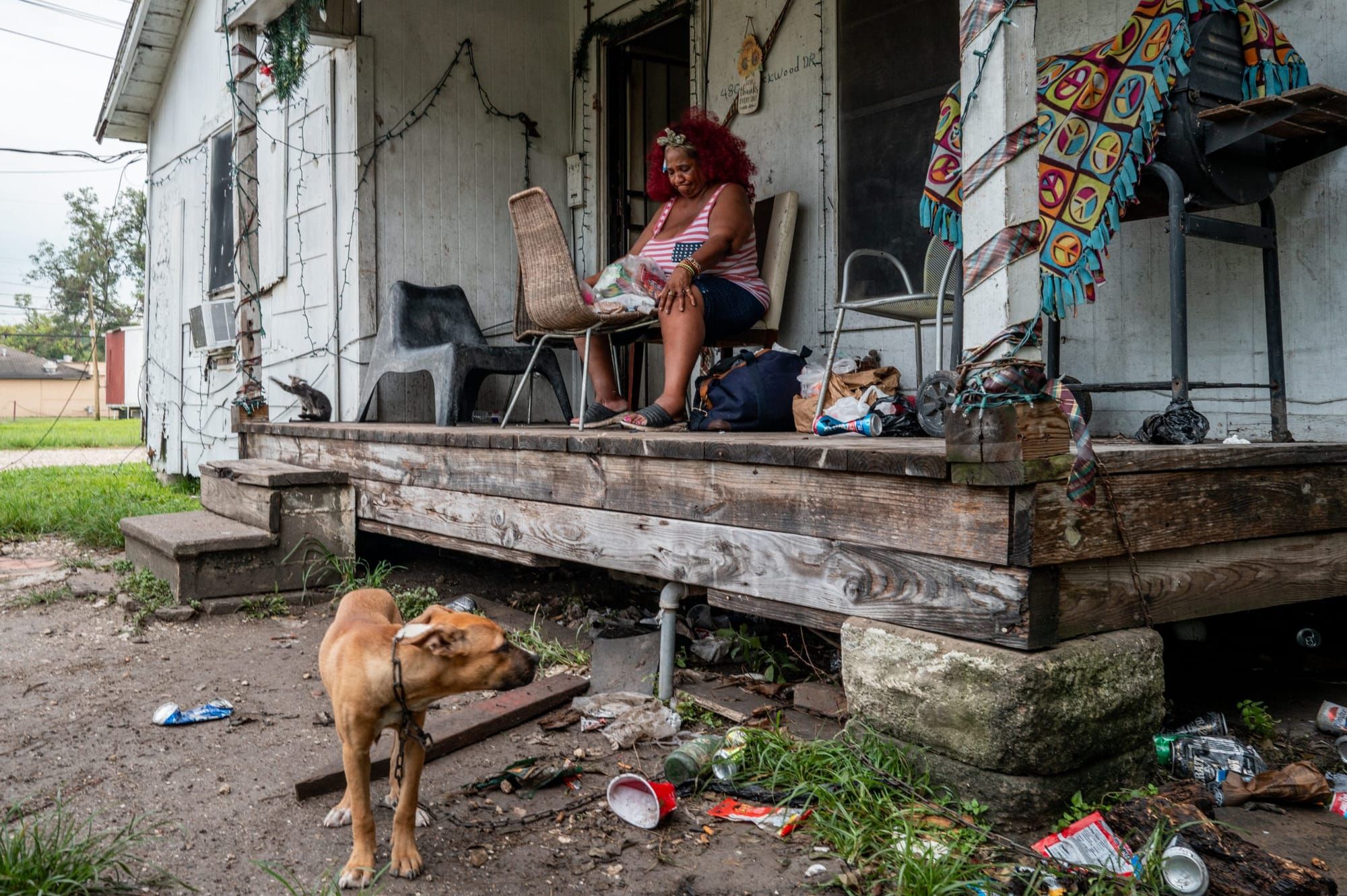 color photograph of a Black woman sitting on a wooden porch outside looking down at a small bag of food in her lap.