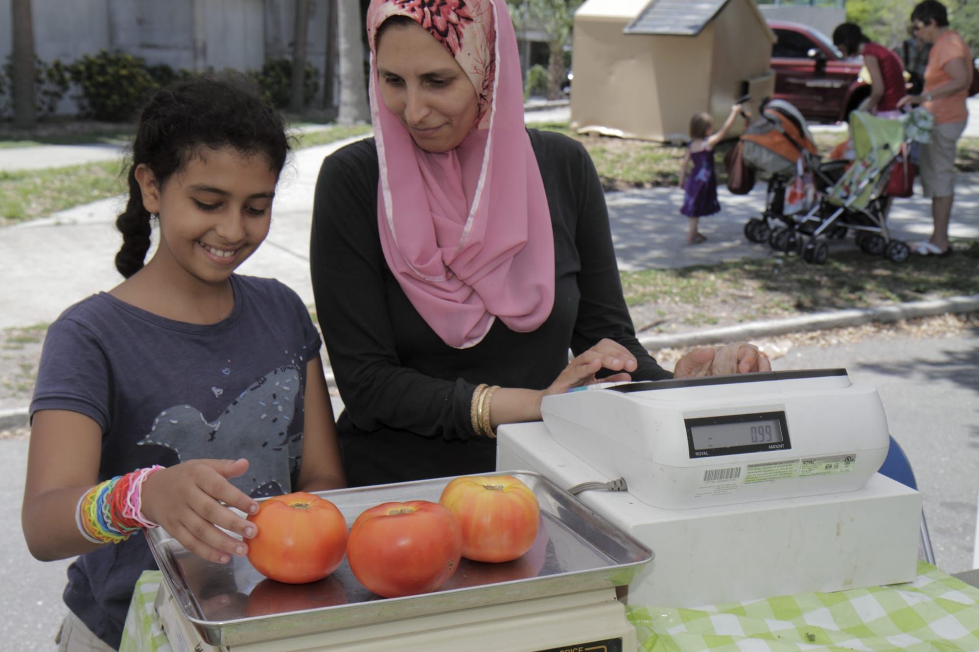 color photograph of a woman wearing a hijab with a young girl at a farmer's market weighing three tomatoes on a digital scale