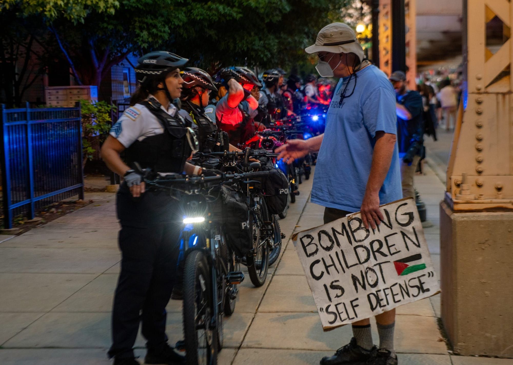photo of a person in a blue shirt and tan bucket hat holding a handmade sign reading, "bombing children is not self defense"