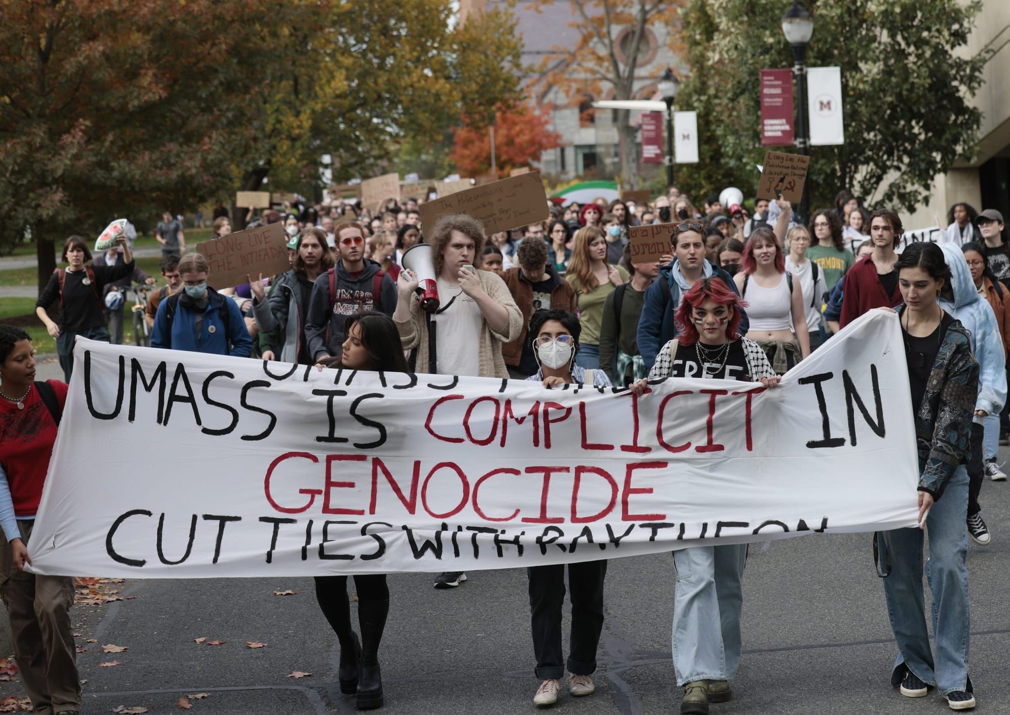 color photograph of an outdoor protest. A group of people walk down the middle of a street holding a banner that reads "UMass