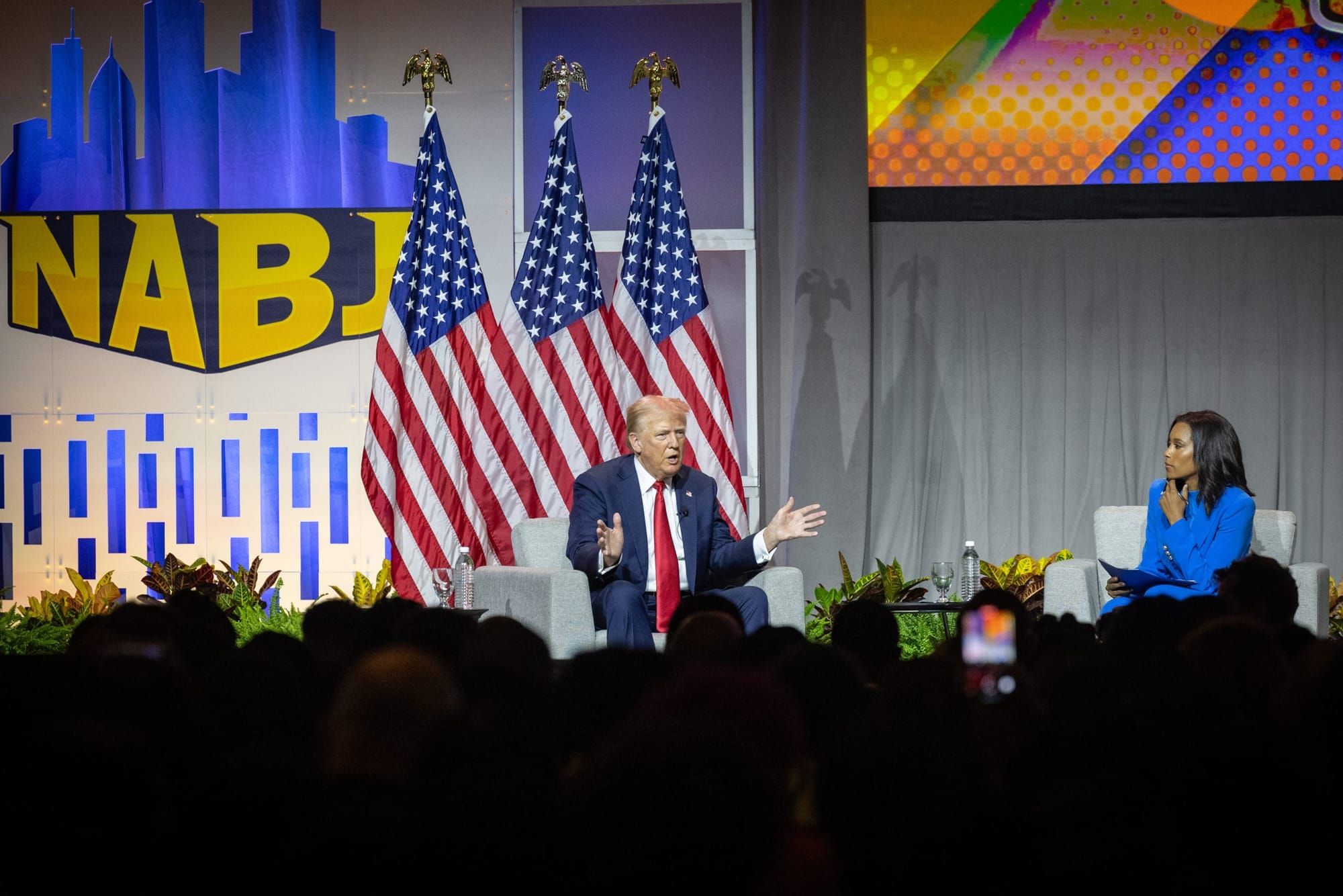 color photograph of Donald Trump sitting on stage at the NABJ convention. three American flags are behind him, and the dark o