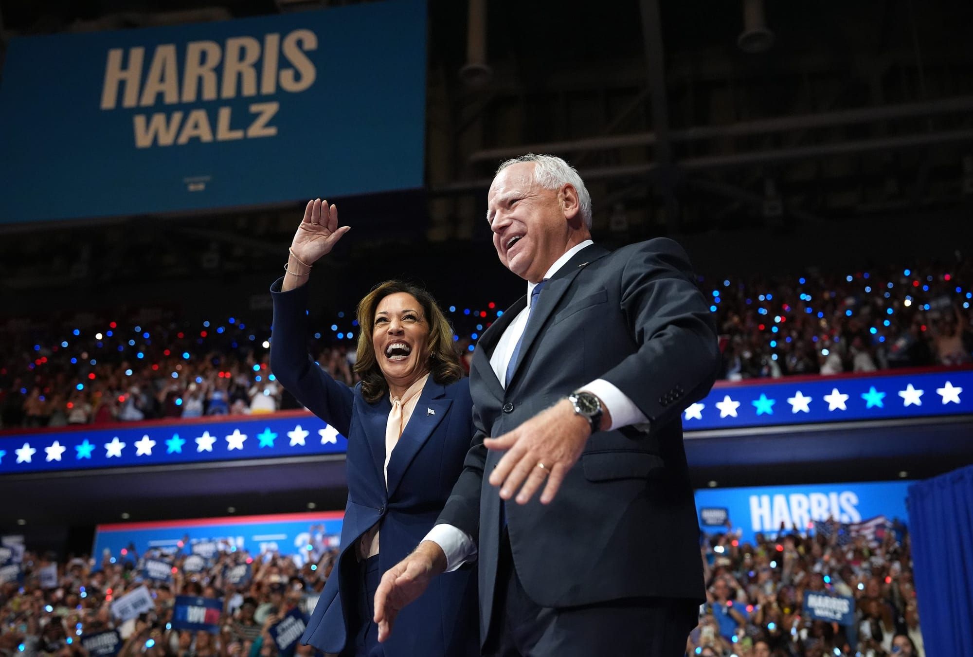 color photograph of Kamala Harris and Tim Walz on stage with their campaign logo behind them hanging over a dense crowd