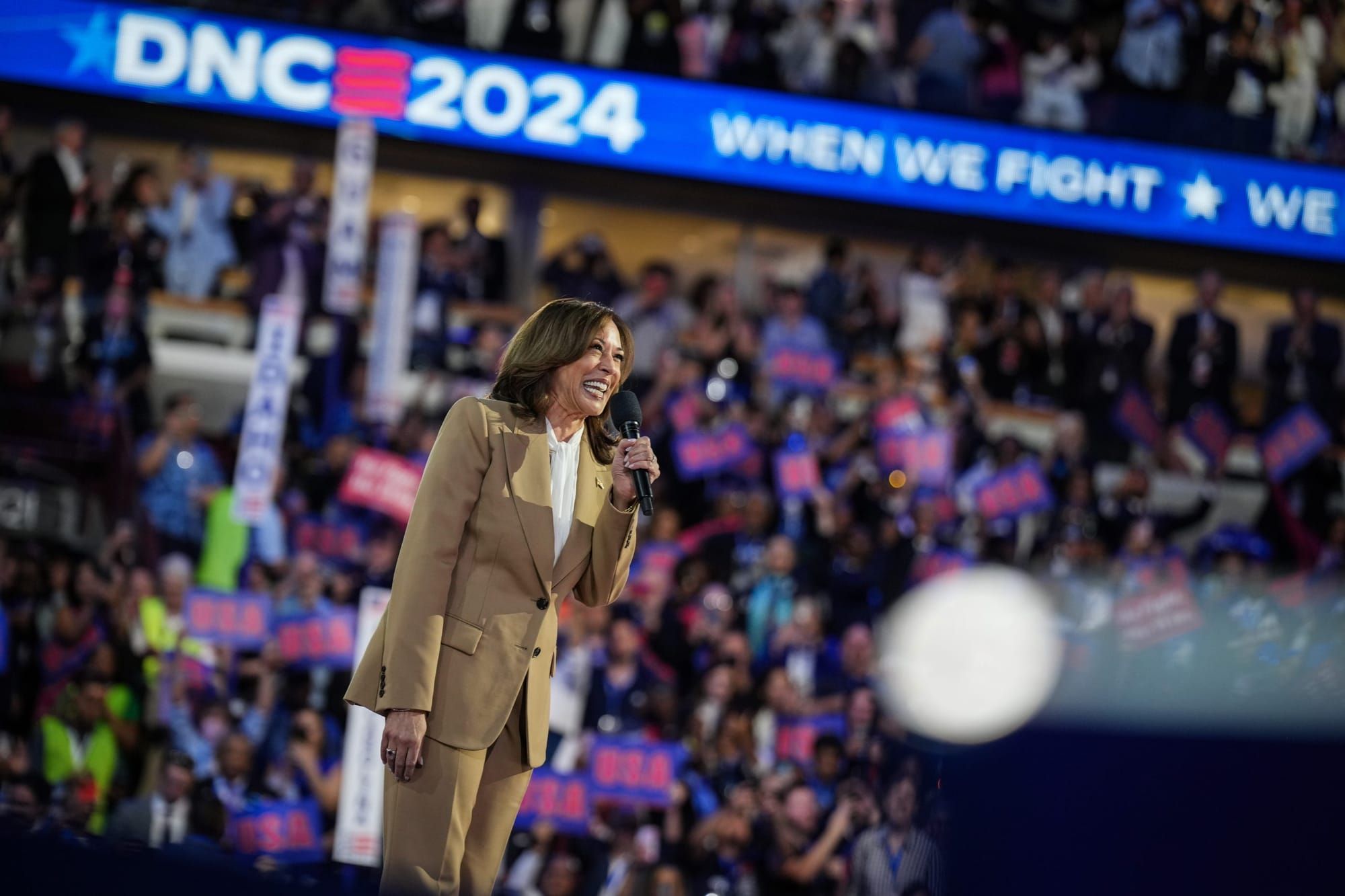 color photograph of Kamala Harris standing on stage at the DNC with a digital banner running along the mezzanine reading "DNC