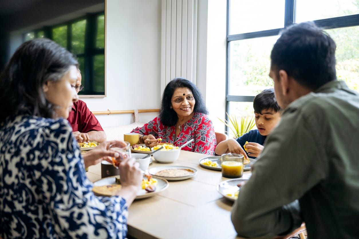 color photograph of a multigenerational Indian family sitting at a kitchen table. Relaxed adults and children look at each ot