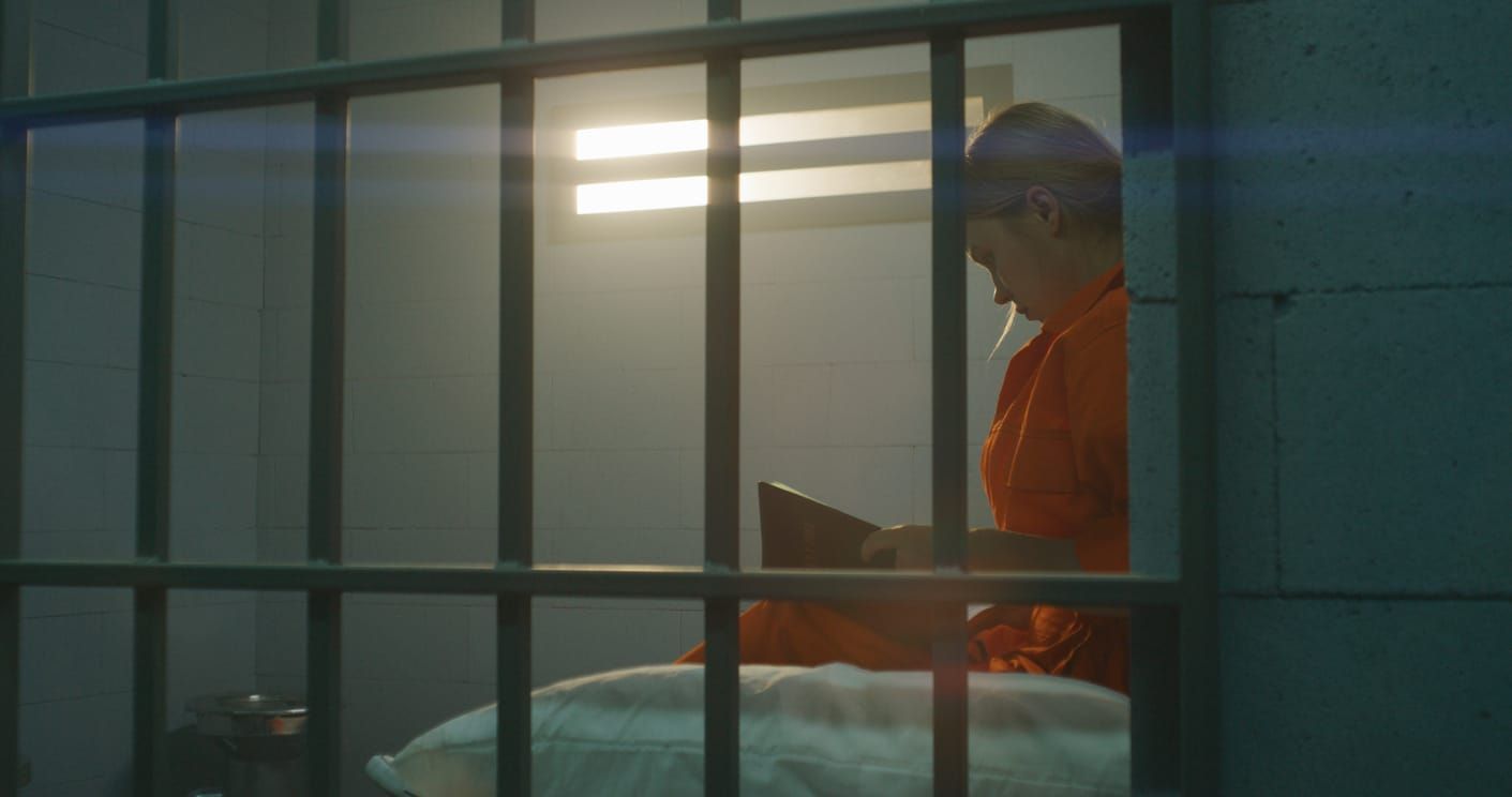 color photo of a woman in an orange prison jumpsuit sitting on a bed behind bars and reading