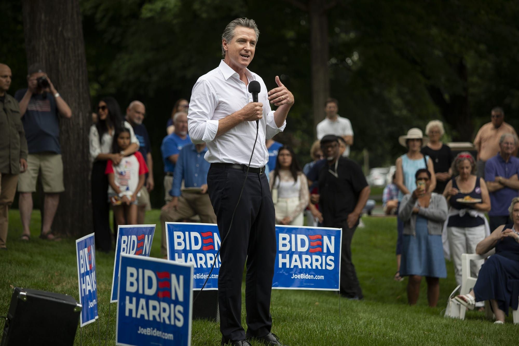 Gov. Gavin Newsom stands on a lawn, holding a microphone and surrounded by "Biden/Harris" campaign signs