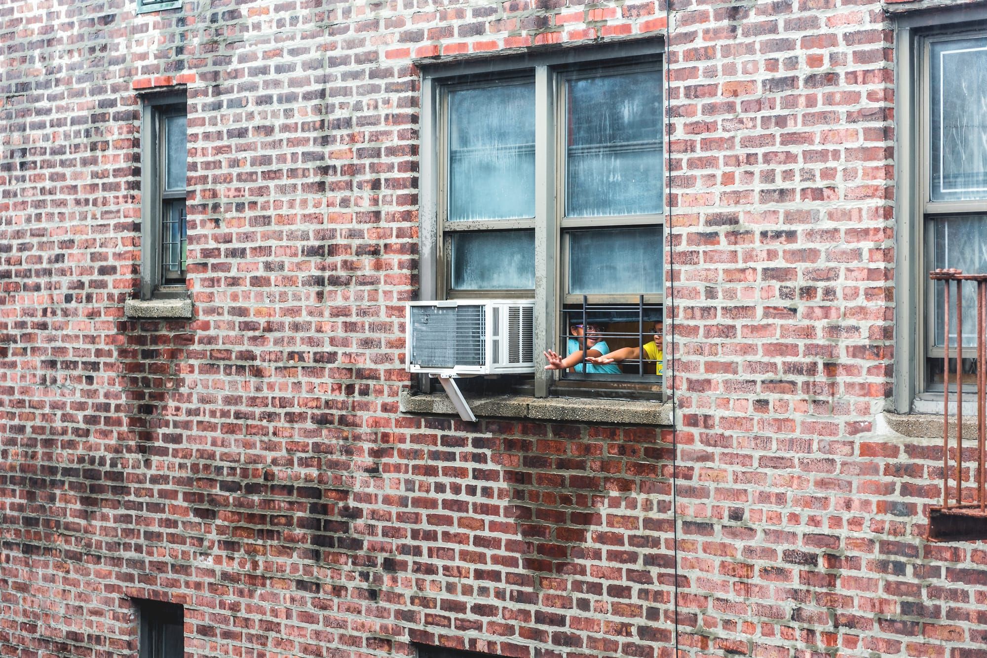 Children stick their hands out of a window next to an air conditioning unit in a brick condo building.