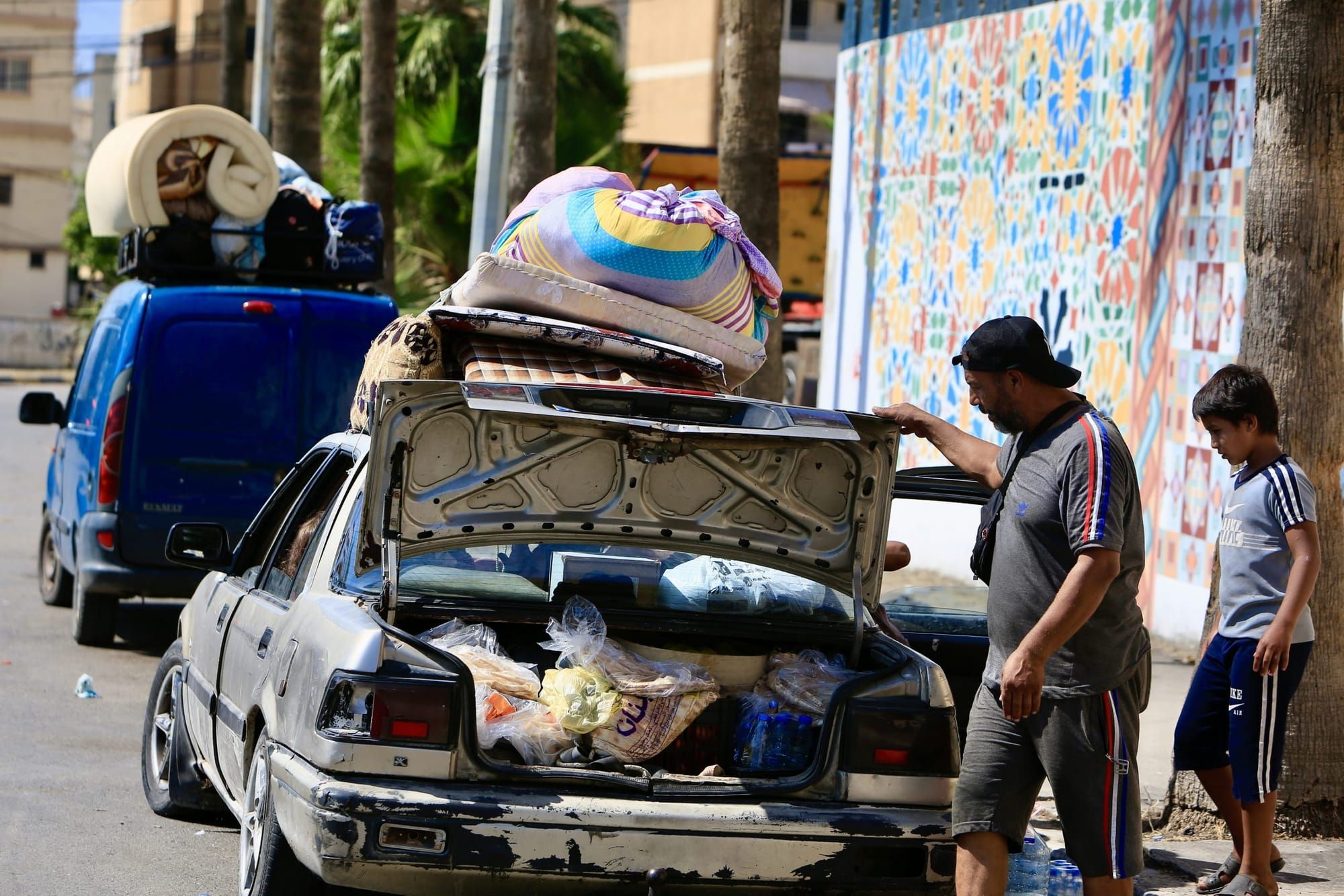A man and boy look at items packed into, and overflowing, the trunk of a car parked on the street.