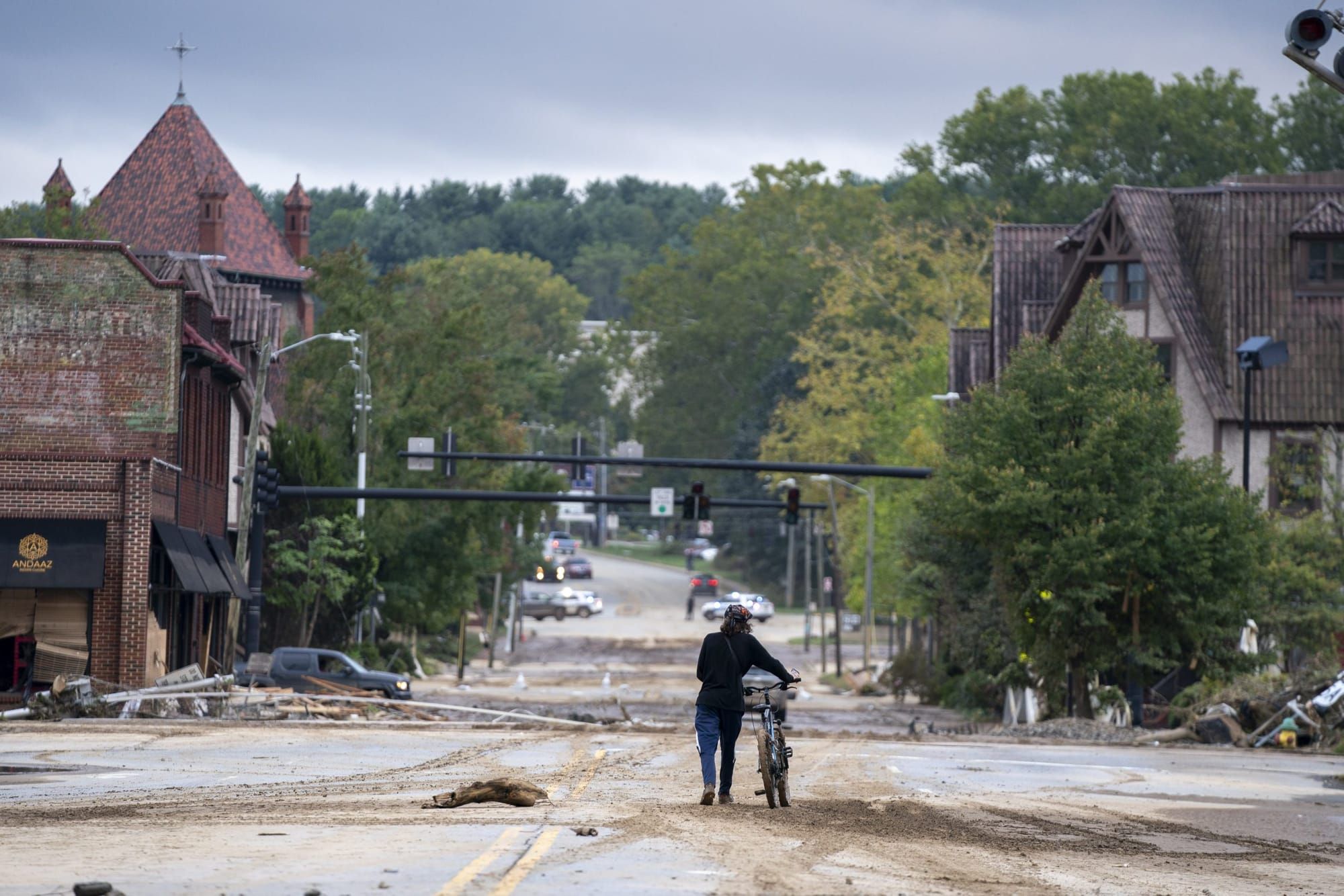 A person walks beside a bicycle thorough a muddy street, buildings on either side show signs of damage