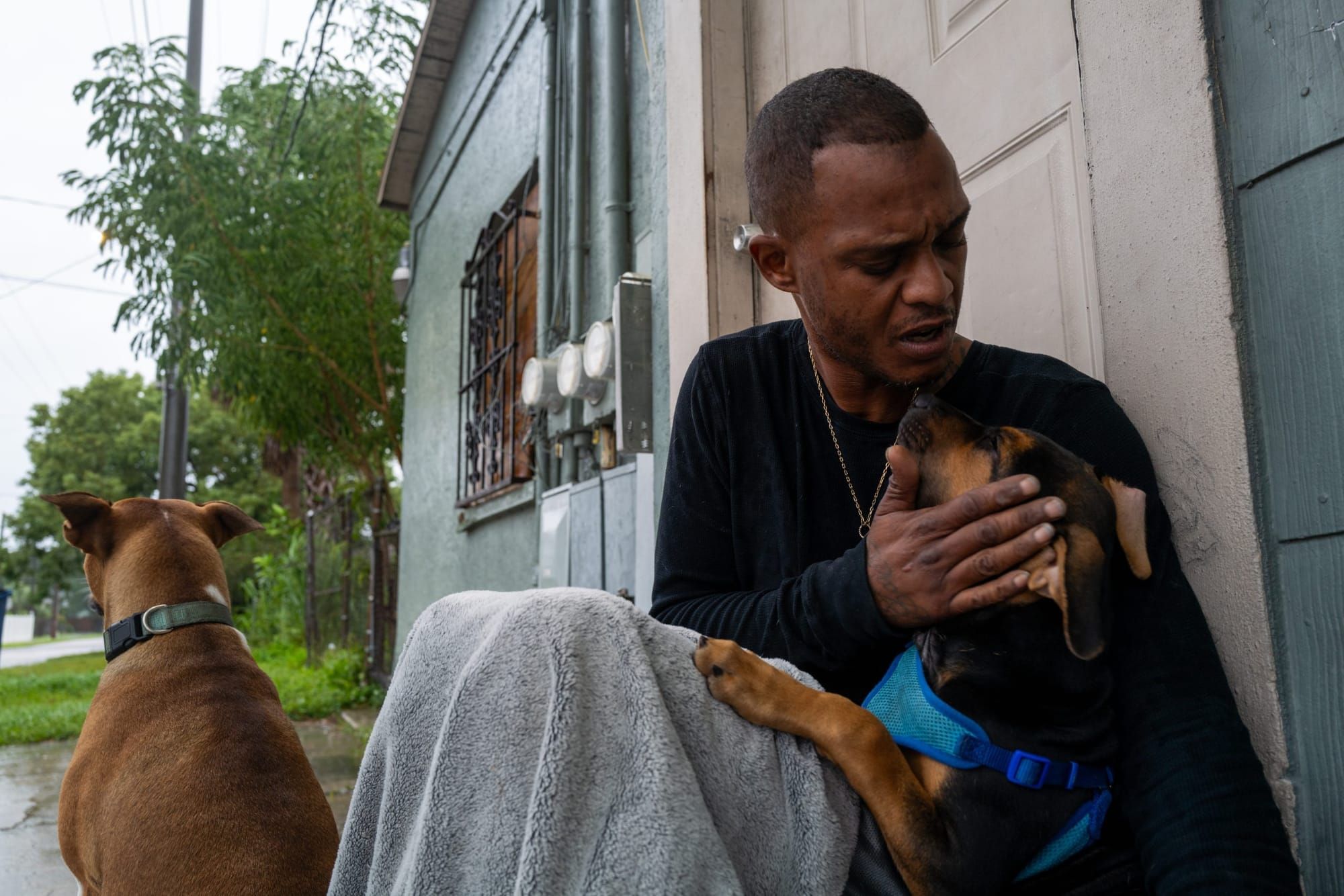 A man sits on the stoop in the back of a building, petting one of the two dogs that sit next to him.