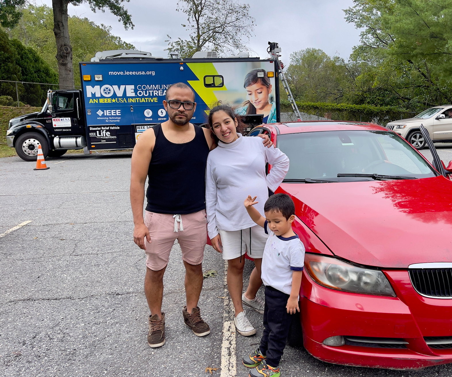 A man, woman and child smile at the camera as they stand next to a red car in a parking lot.