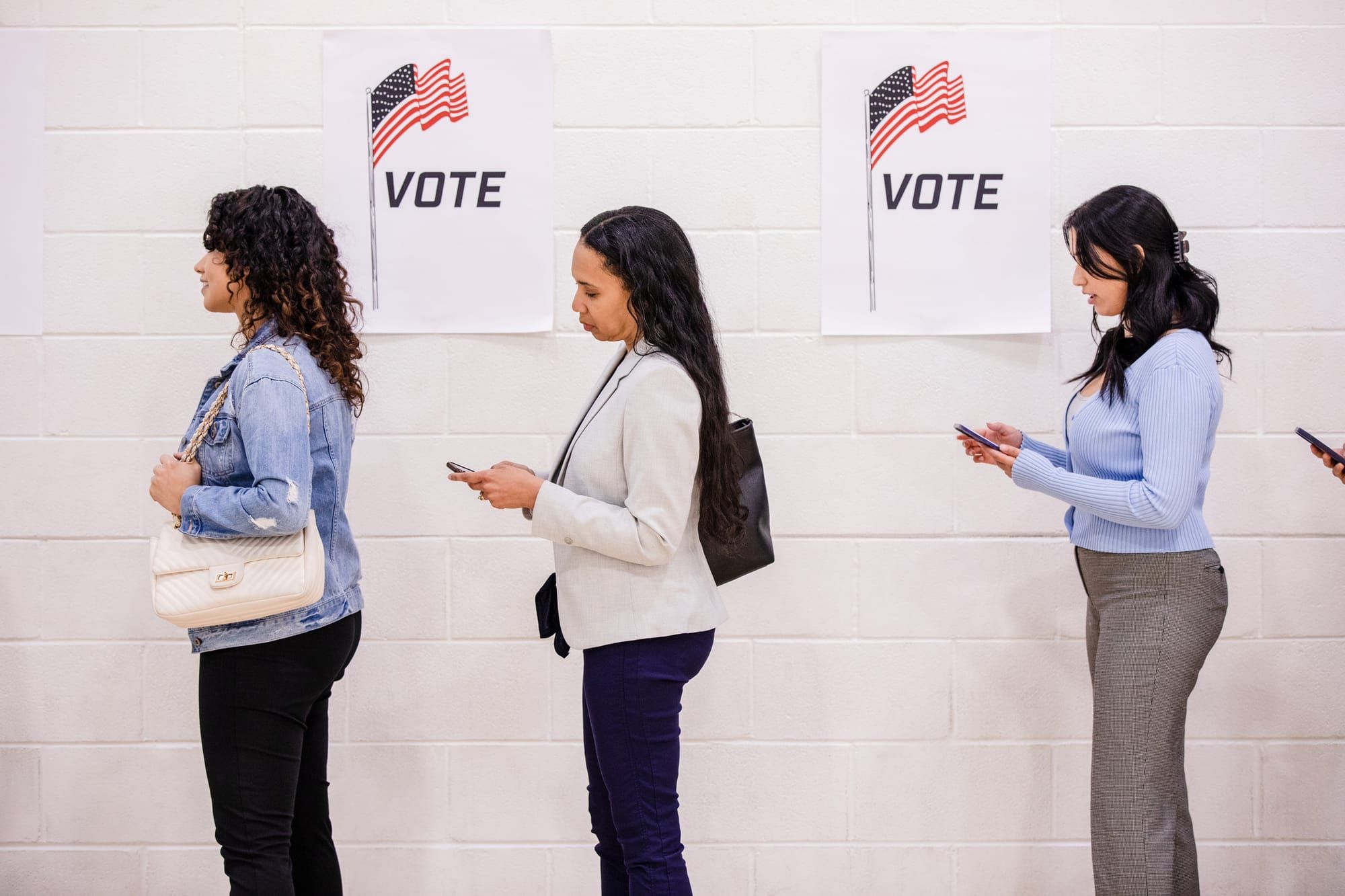 Young women look at their phones while standing in line to vote.