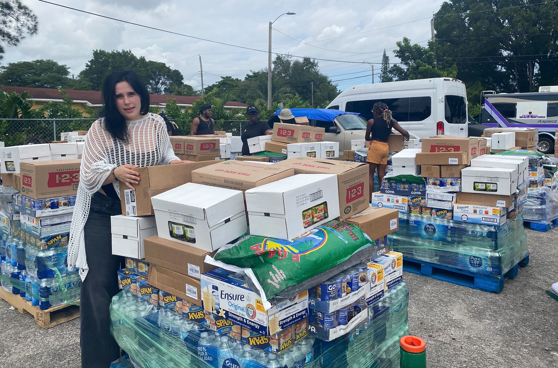 A woman stands next to boxes and crates of water and nonperishable food items.