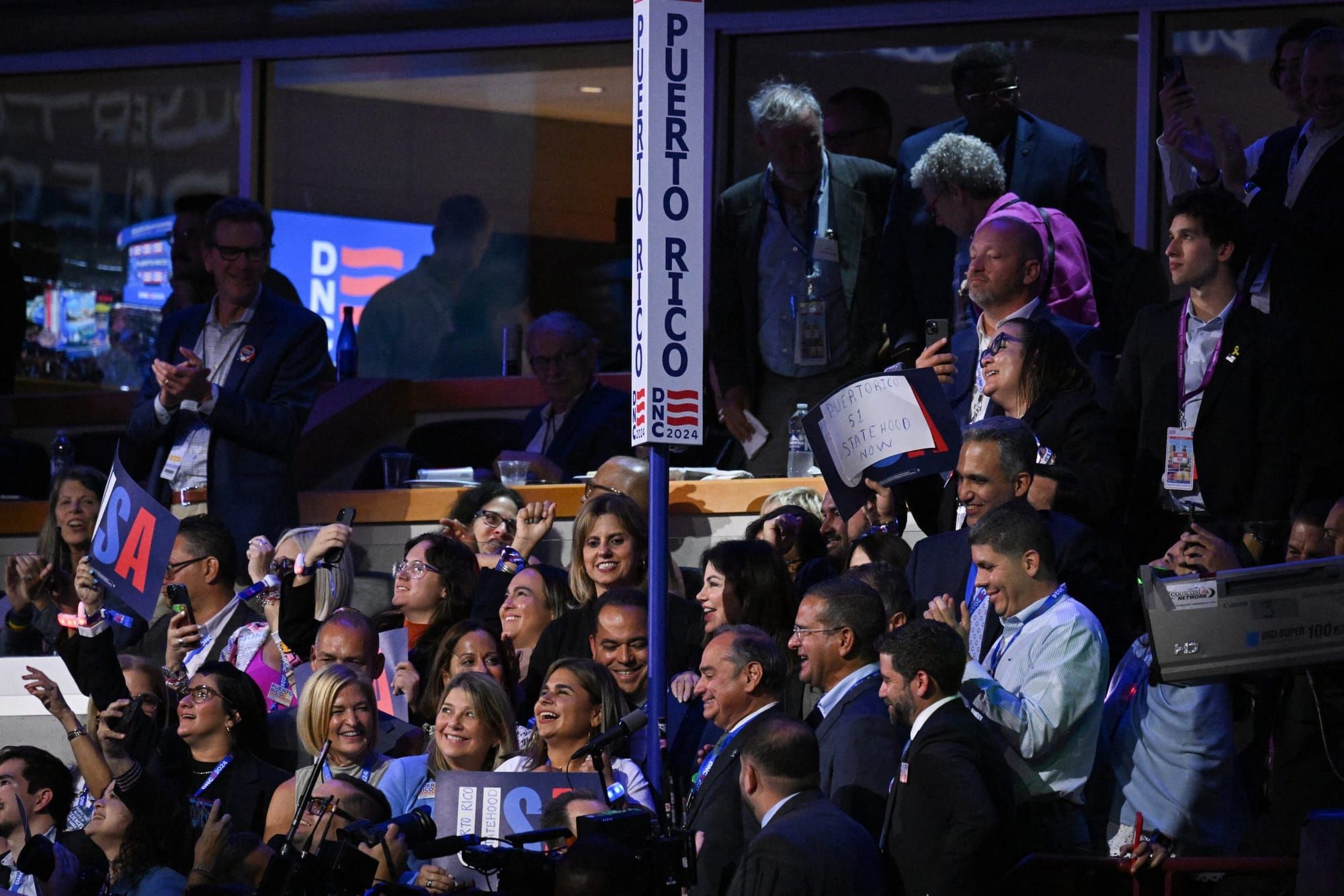 Delegates dressed in suits sit in stadium seating near a vertical "Puerto Rico" sign