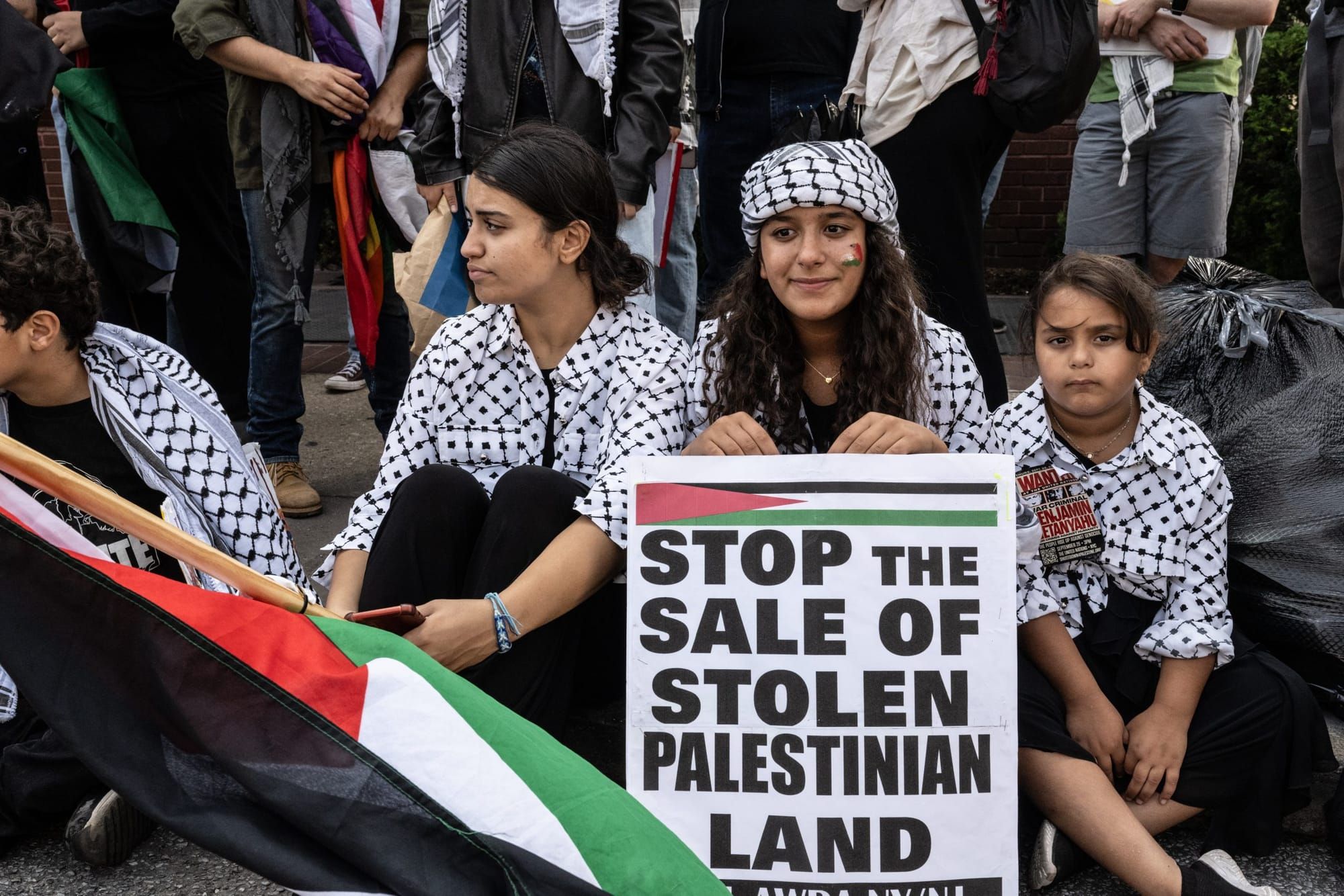 Protesters sit on a curb holding a Palestinian flag, each wearing a keffiyeh. One of the protesters holds a sign that says, "