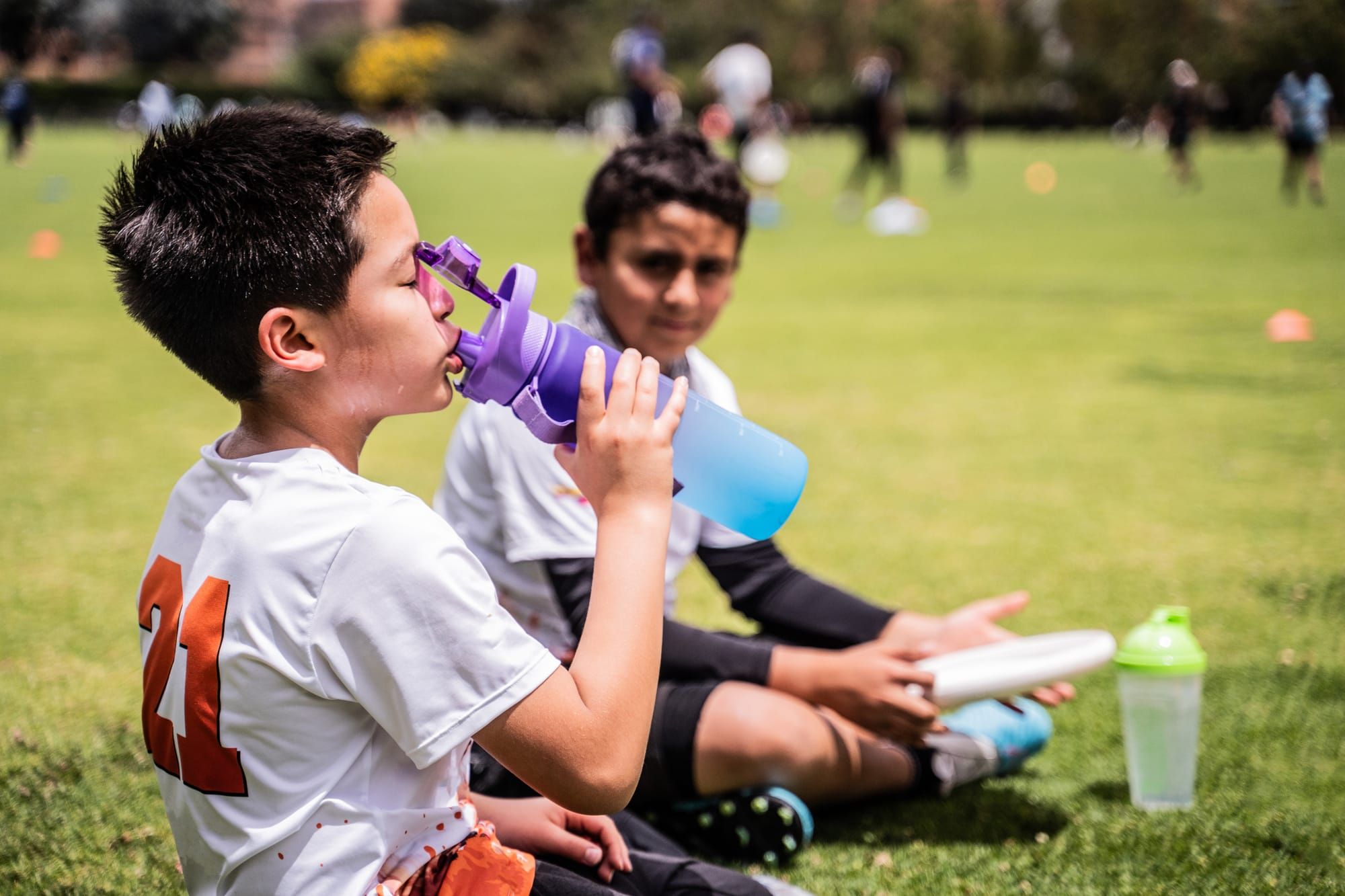 Two boys sit on a soccer field, one watching the other drink water from a water bottle.