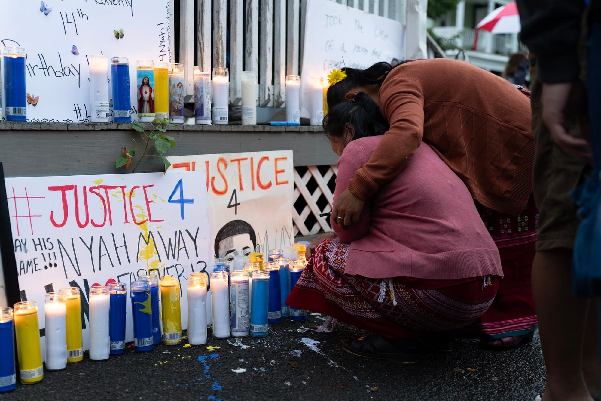 woman sits in front of posters that say Justice 4 Nyah Mway and candles, while another woman puts her arm around her