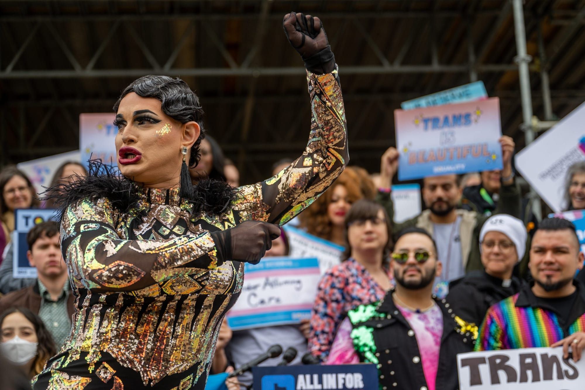 drag performer poses, with protesters carrying signs in support of trans rights in the background