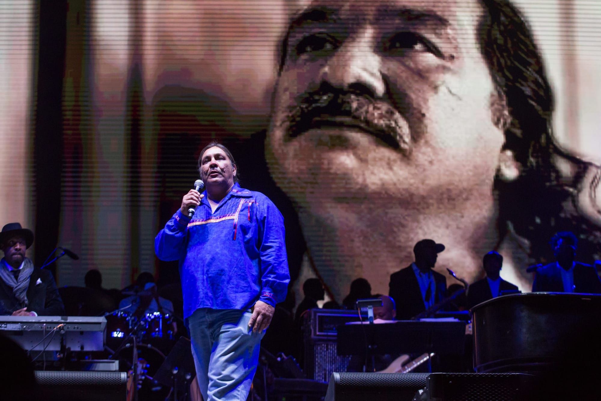 man in spotlight talking in microphone, with image of Leonard Peltier's face on screen in the background