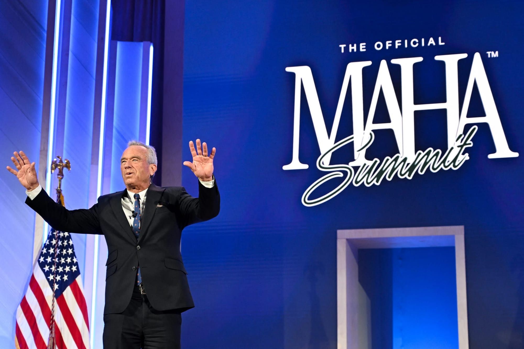 Robert F. Kennedy Jr. on stage waving to the crowd with a MAHA summit sign on a blue background