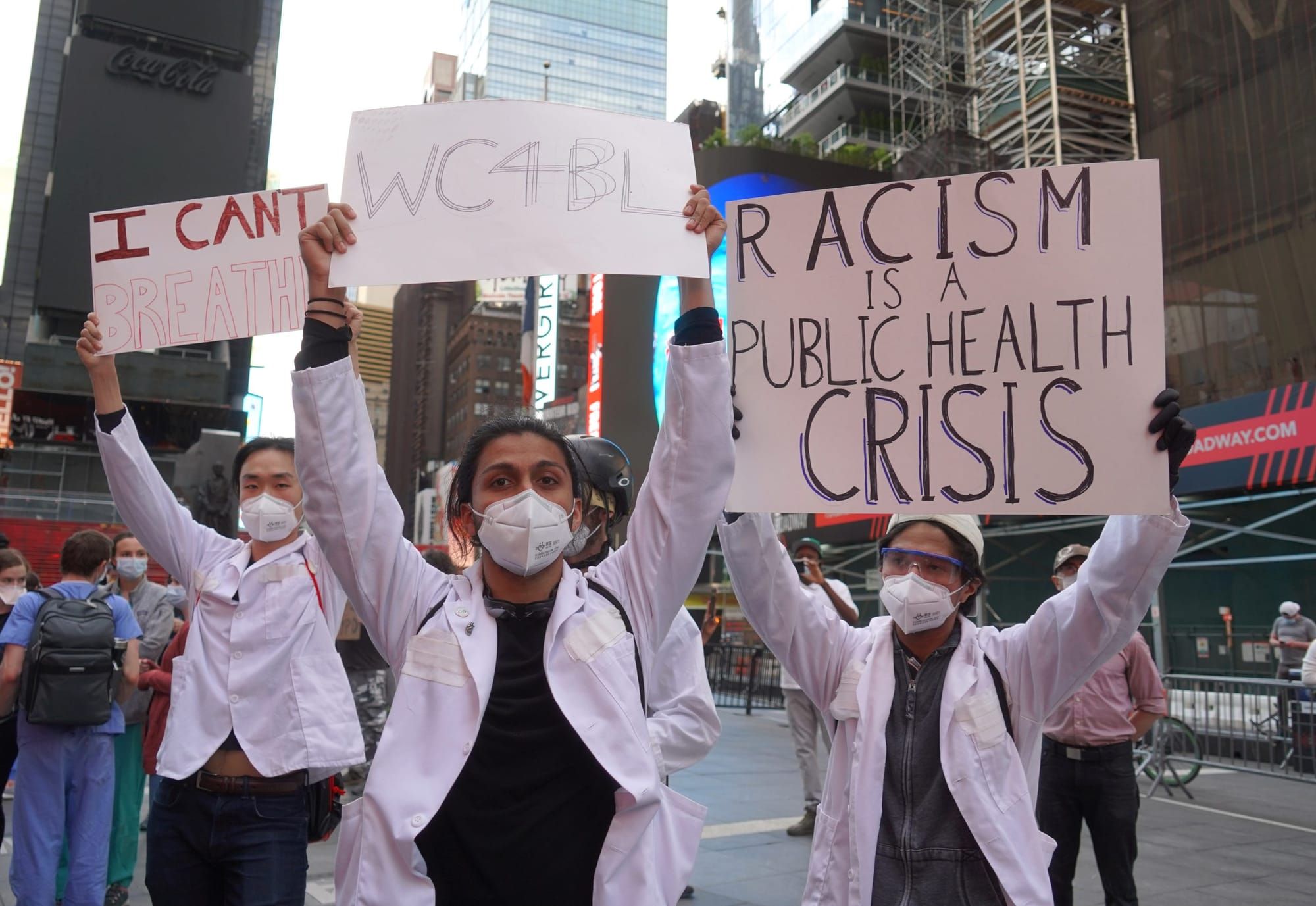 protesters dressed in white coats and masks holding signs up that read "I can't breathe" and "Racism is a public health crisi