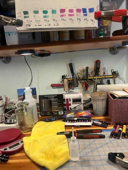 My workbench converted to marker refill station. There is a pair of linesman pliers, a squishy plastic bottle that holds 66ml of water, a yellow low lint rag, and a kitchen quality scale set to zero grams. A pile of different colored alcohol markers has been laid out, sorted by prismacolor identifier.