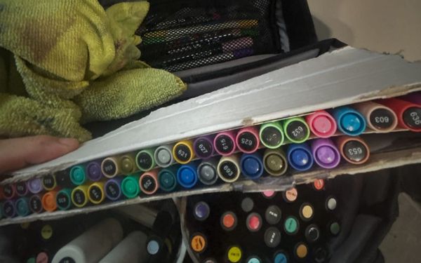 A converted photo gear bag stuffed with alcohol markers and acrylic paint pens. A box of pens is lifted towards the camera.